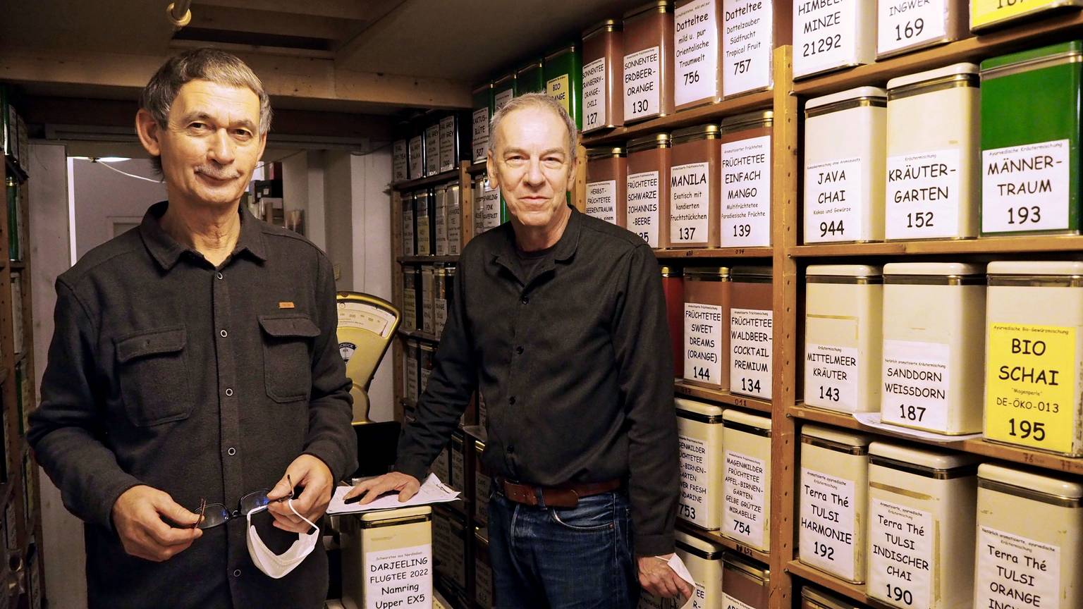 Zwei Männer um die 60 stehen neben der Wand mit den unterschiedlichen Teesorten. Sie tragen schwarze Hemden und Jeans.