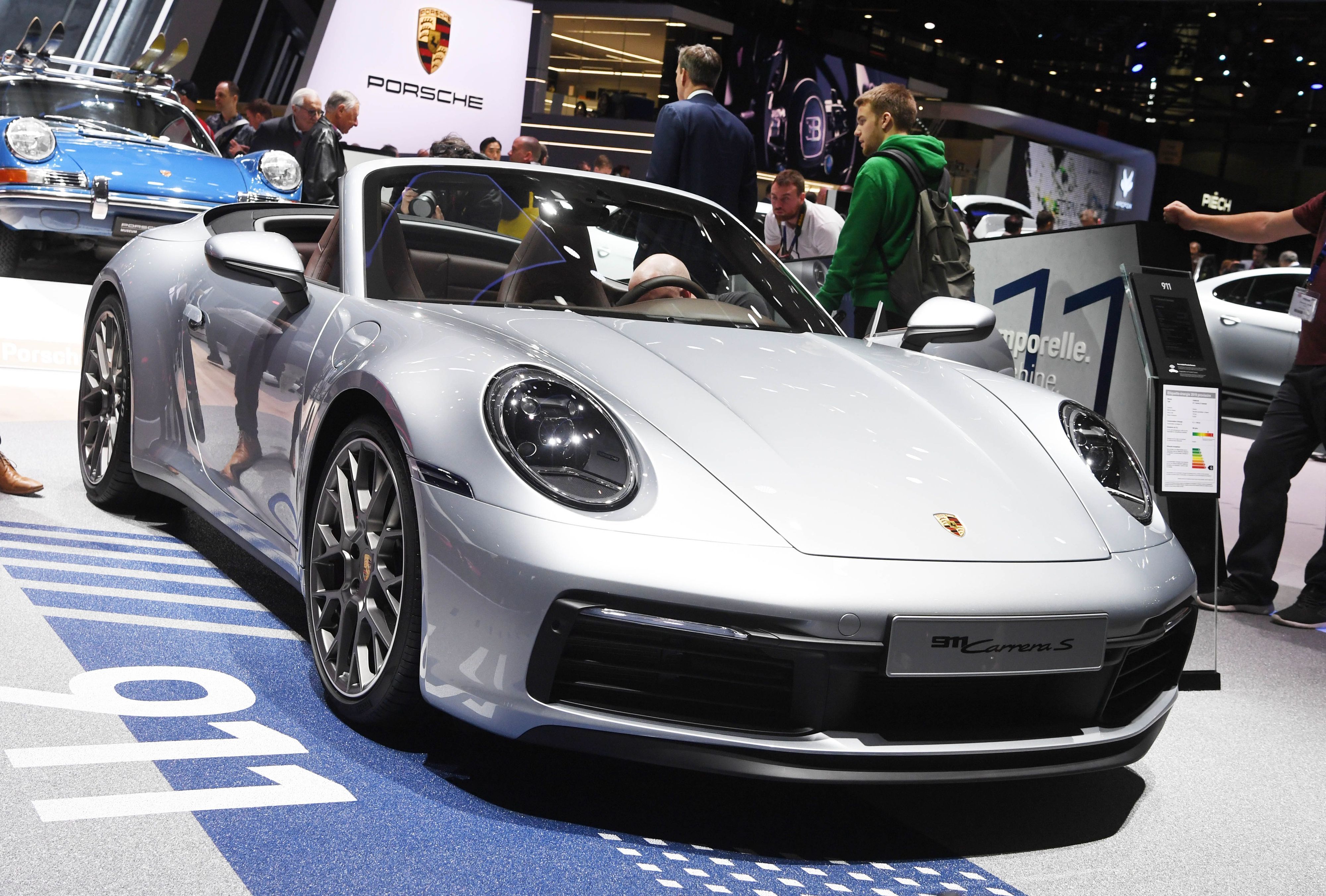 Auf dem Genfer Autosalon wird ein Porsche 911 Cabriolet präsentiert. Es it ein silberner Sportwagen mit dem Porsche-Logo.