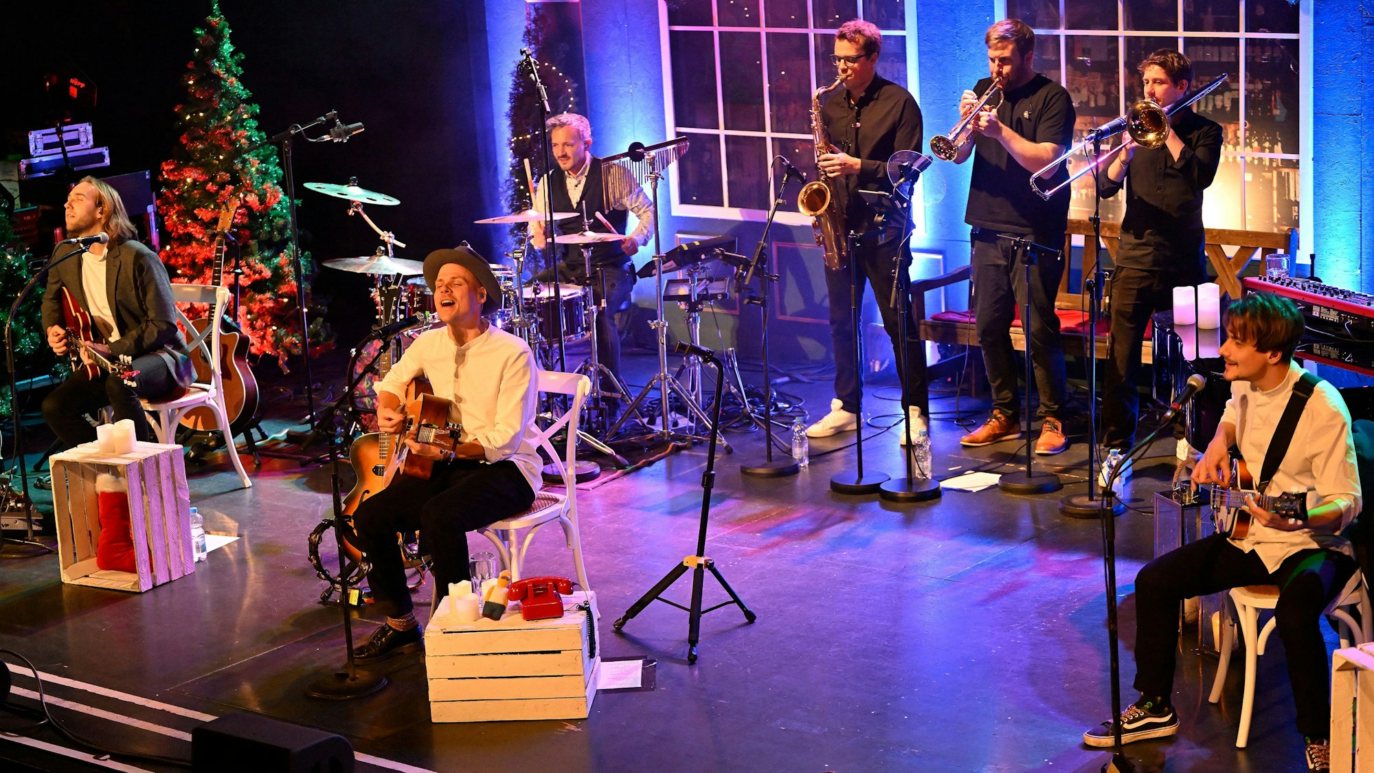 Musiker spielen zwischen Weihnachtsbäumen und großen Fenster in violettem bis warmweißen Licht.