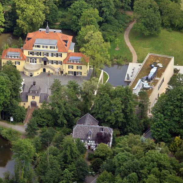 Eine Luftaufnahme von Schloss Eulenbroich inmitten von Waldgebiet und einer Wiesenfläche, die Platz für einen Spielpatz bieten könnte.