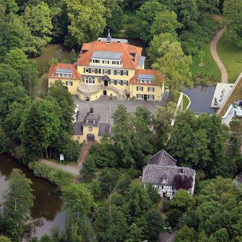 Eine Luftaufnahme von Schloss Eulenbroich inmitten von Waldgebiet und einer Wiesenfläche, die Platz für einen Spielpatz bieten könnte.