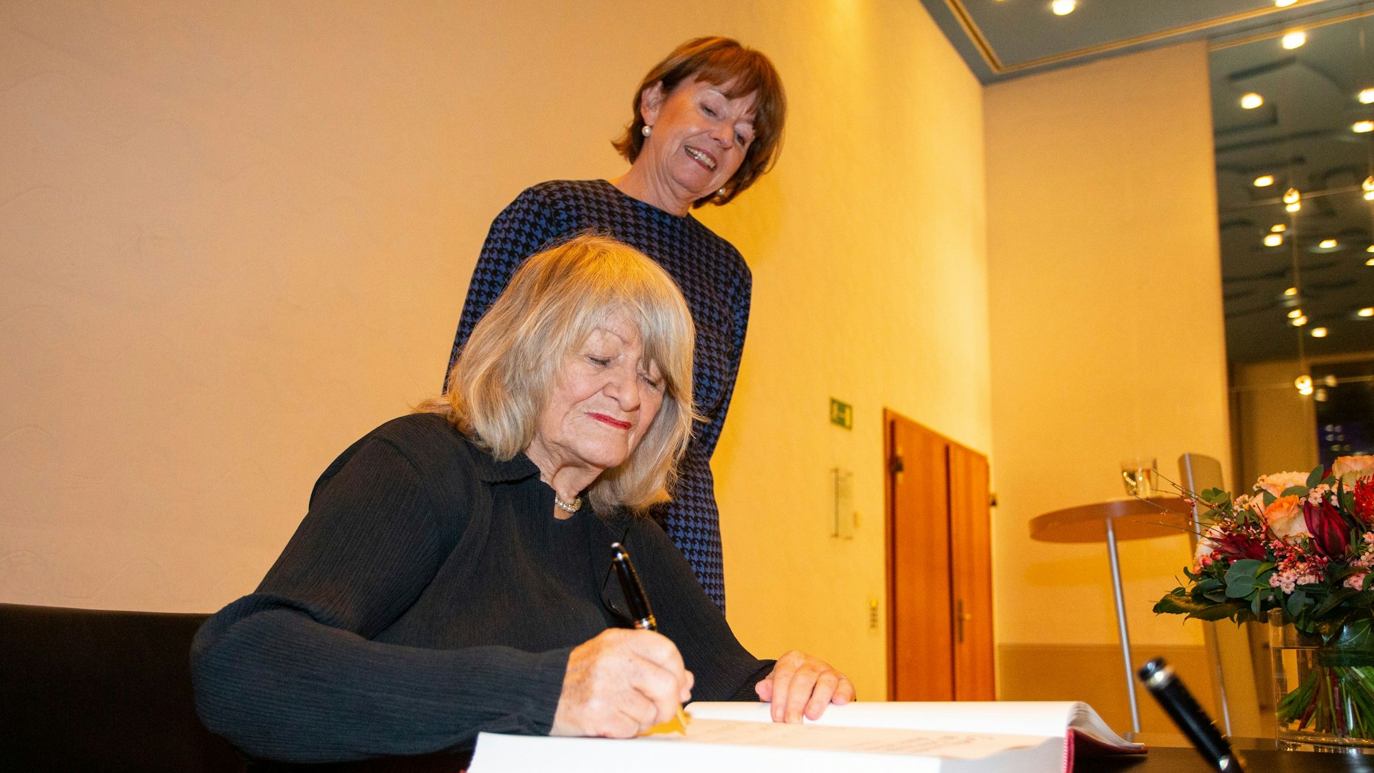 Alice Schwarzer trägt sich ins Gästebuch der Stadt Köln ein. Hinter ihr ist Kölns Oberbürgermeisterin Henriette Reker zu sehen, die ihr über die Schulter schaut.