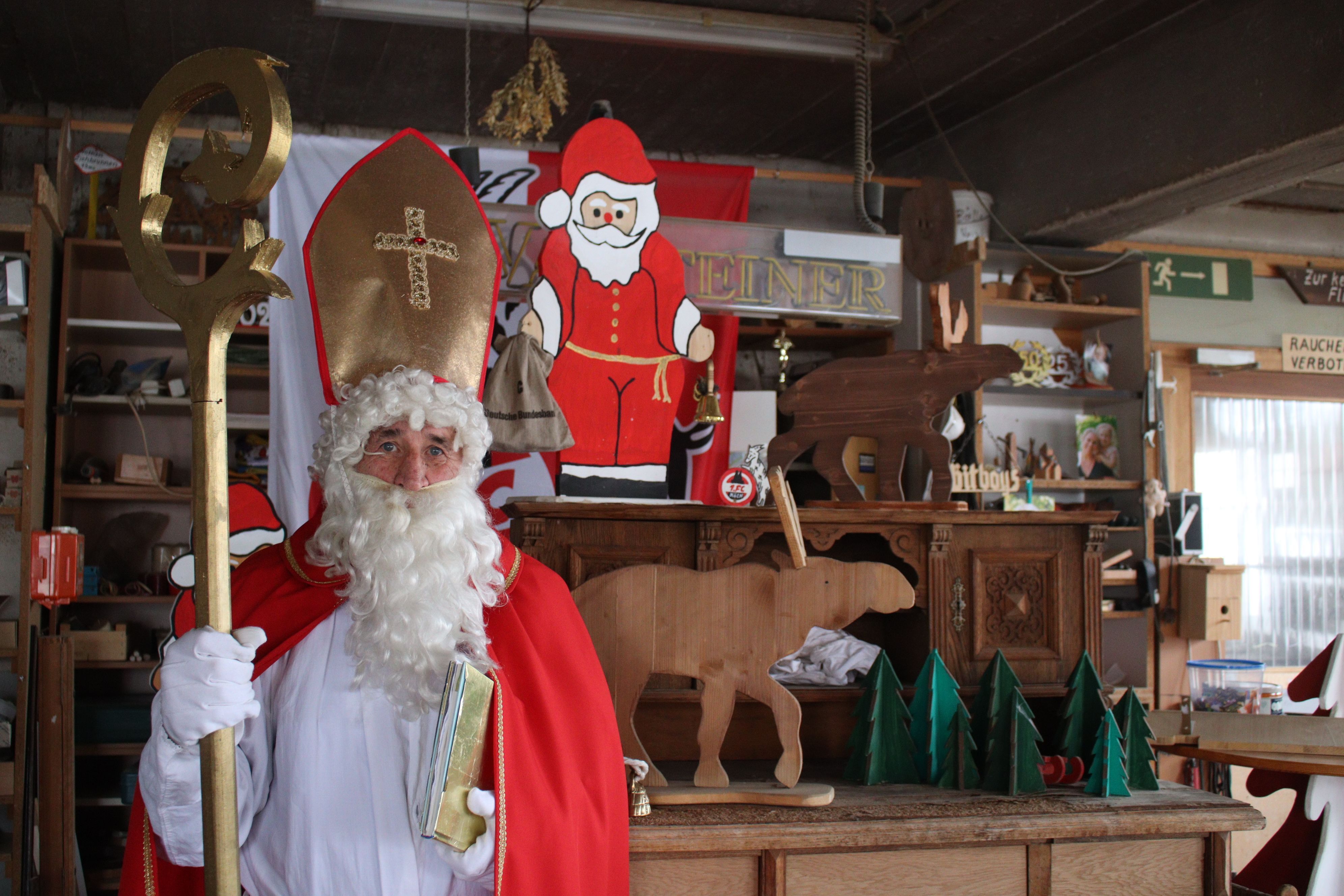 Hans-Peter Lückerath, genannt Hacky, steht im Nikolausgewand mit Stab und goldenem Buch in der Hand in seiner Schreinerwerkstatt in Nöthen. Im Hintergrund befinden sich unter anderem ein Nikolaus, Elche und Weihnachtsbäume aus Holz.