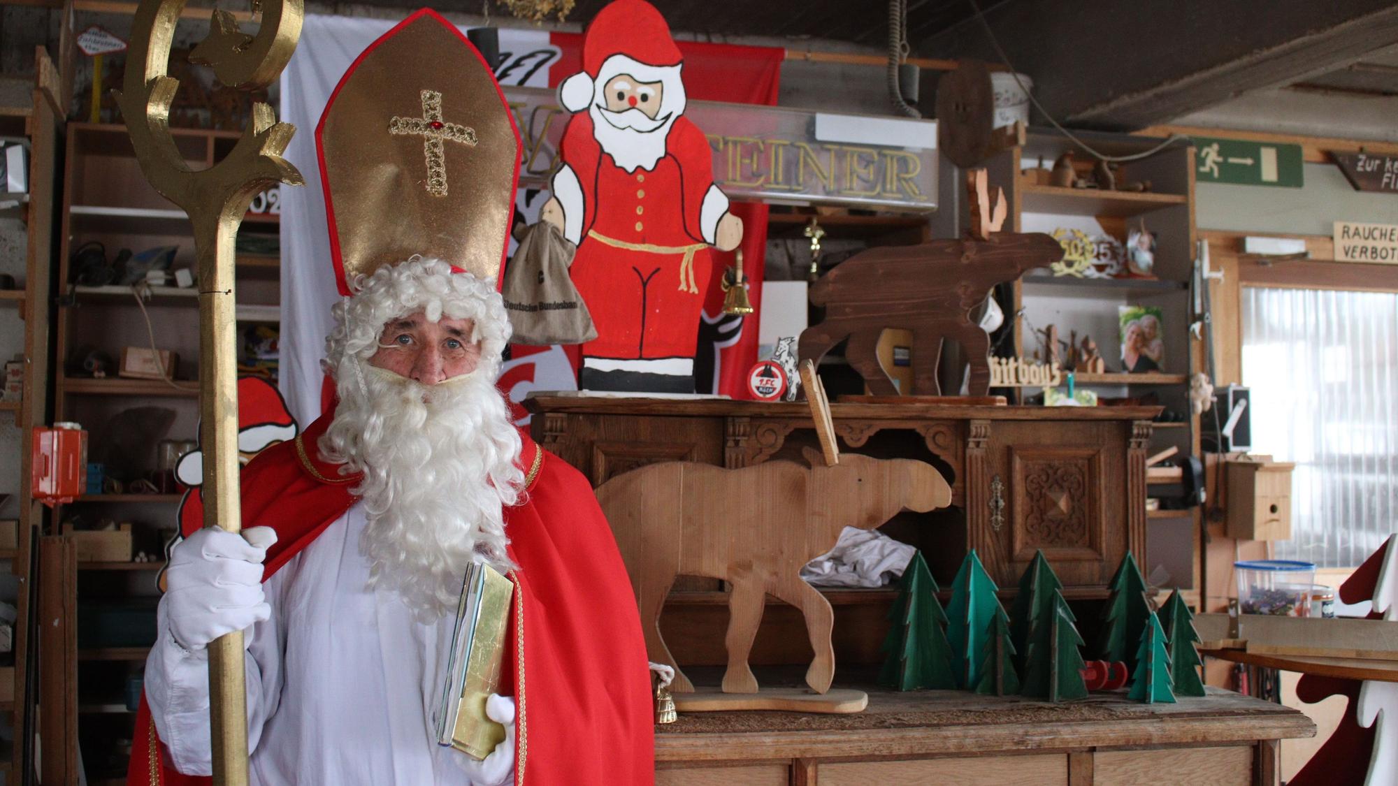 Hans-Peter Lückerath, genannt Hacky, steht im Nikolausgewand mit Stab und goldenem Buch in der Hand in seiner Schreinerwerkstatt in Nöthen. Im Hintergrund befinden sich unter anderem ein Nikolaus, Elche und Weihnachtsbäume aus Holz.