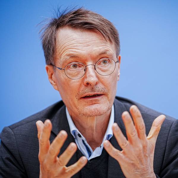 Bundesgesundheitsminister Karl Lauterbach sitzt im Anzug vor blauem Hintergrund auf eine Pressekonferenz. Er hält die Hände offen vor der Brust.