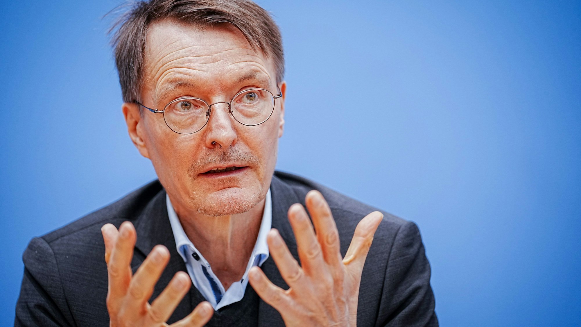 Bundesgesundheitsminister Karl Lauterbach sitzt im Anzug vor blauem Hintergrund auf eine Pressekonferenz. Er hält die Hände offen vor der Brust.