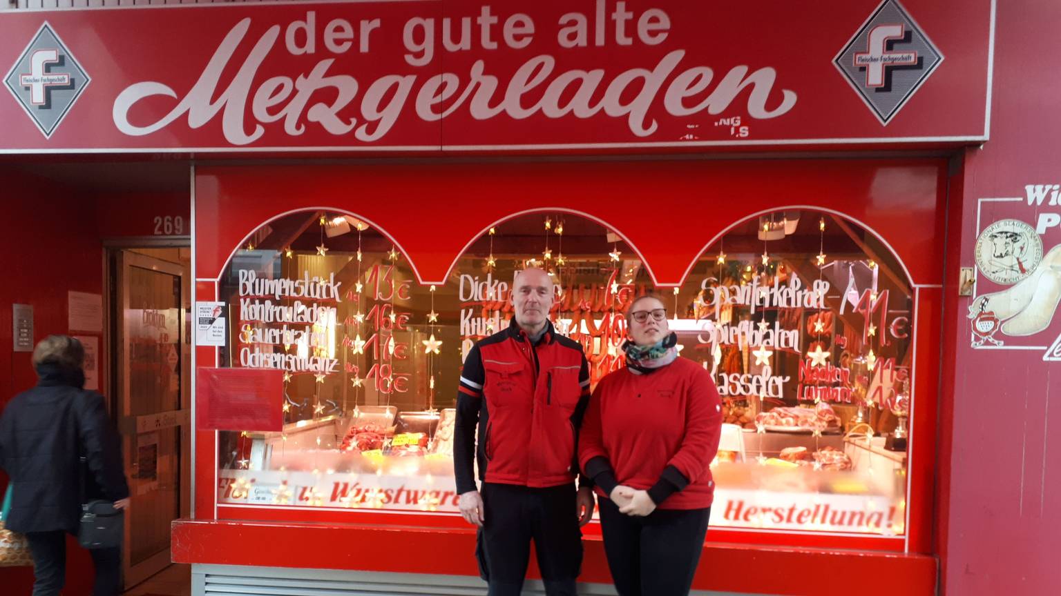 Ein Mann mittleren Alters mit Glatze und eine Frau stehen vor dem in rot gehaltenen Ladenlokal der Metzgerei, die ihnen gehört.