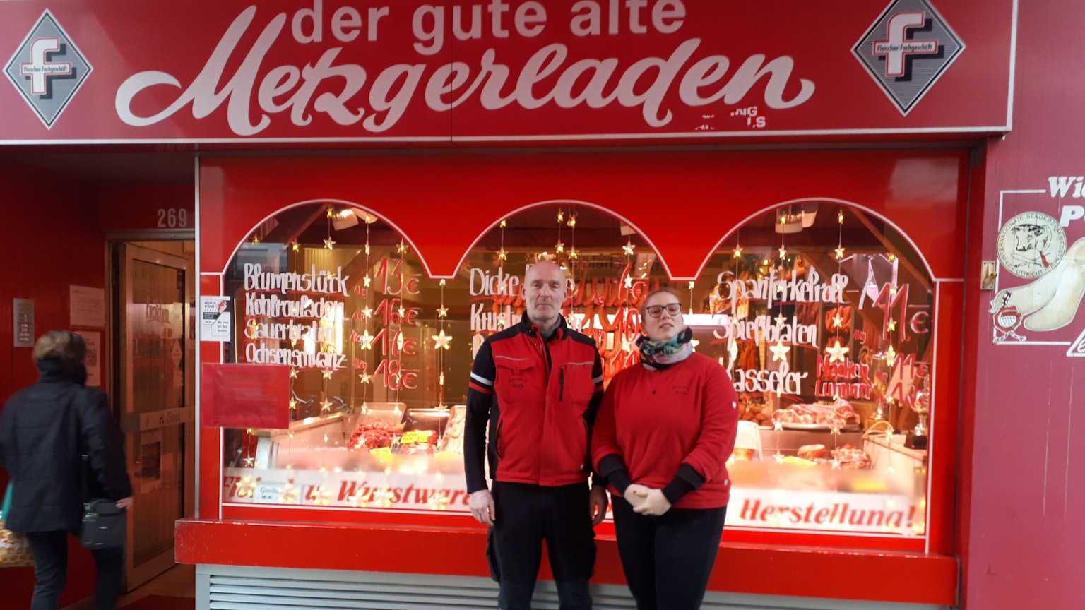 Ein Mann mittleren Alters mit Glatze und eine Frau stehen vor dem in rot gehaltenen Ladenlokal der Metzgerei, die ihnen gehört.