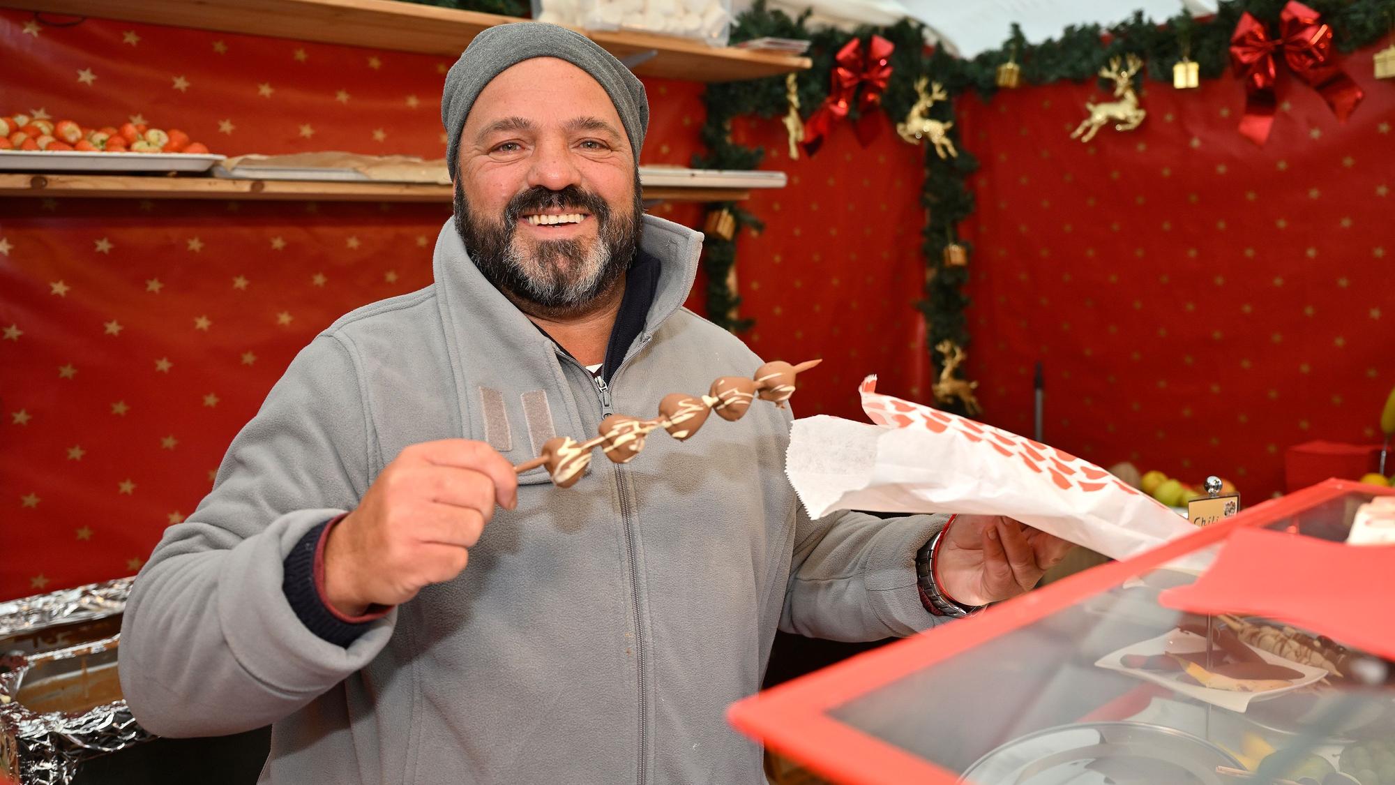 STefan Reinhard, ein Mann in grauen Klamotten, Mütze und mit Bart, zeigt einen Spieß mit mit Schokolade überzogenen Früchten.