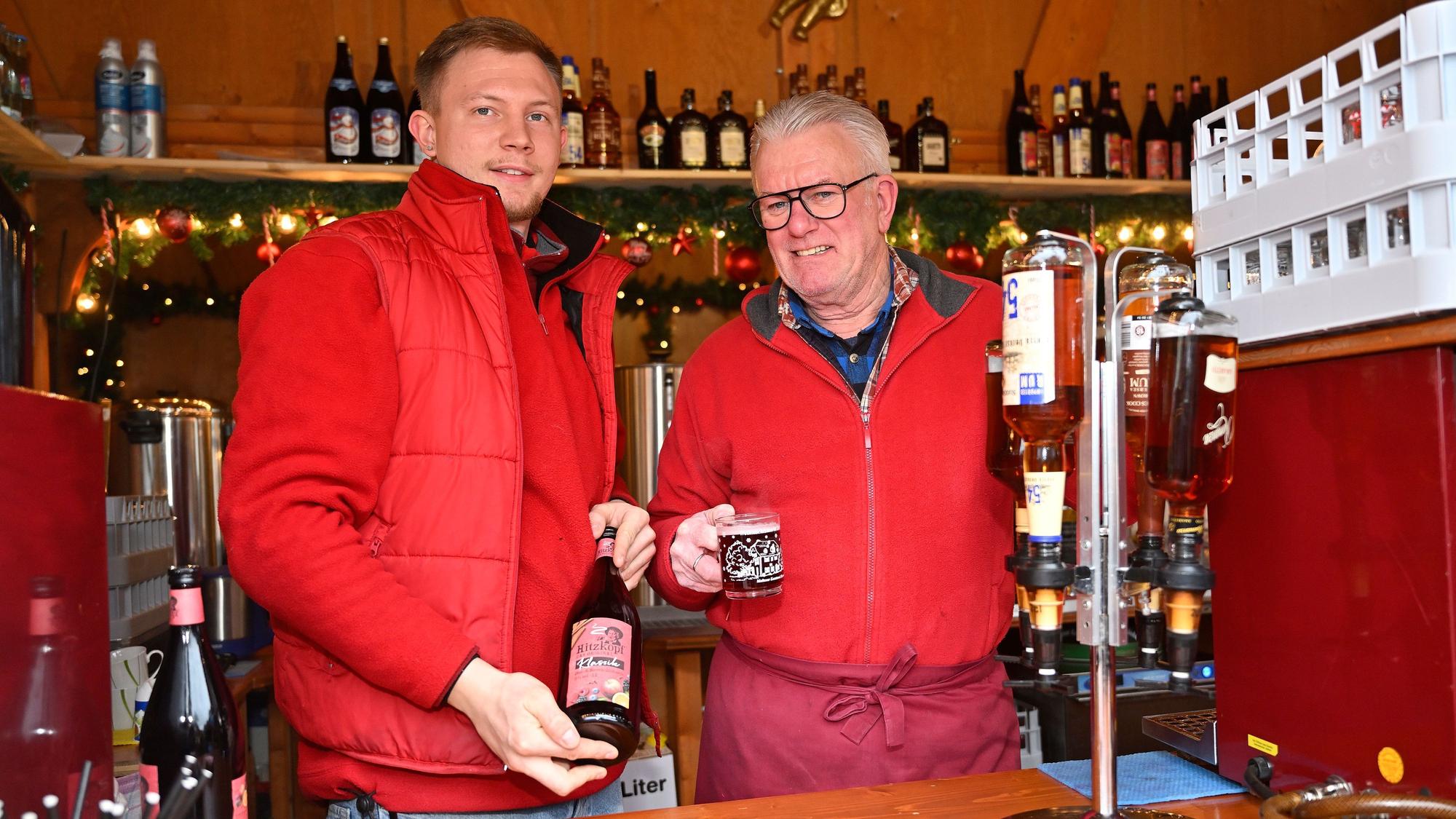 Alexander Timm und Peter Germerodt stehen in ihrer Glühweinbude und zeigen eine Tasse mit roten Glühwein und eine volle Flasche vor.