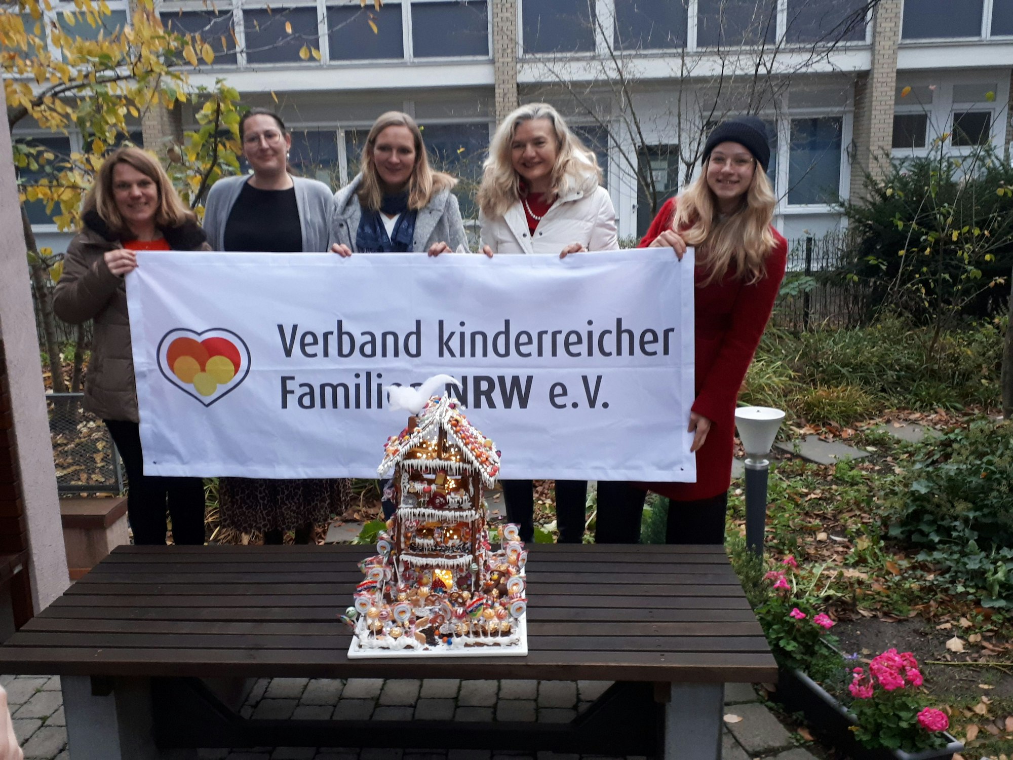 Fünf Frauen halten ein Transparent hoch mit dem Schriftzug Verband kinderreicher Familien NRW e.V. Auf einer Bank davor steht ein liebevoll verziertes vier Etagen hohes Lebkuchenhaus.