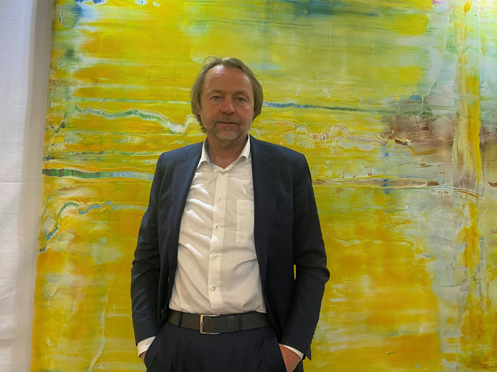 Ein Mann in einem dunklen Anzug und weißem Hemd steht vor einem großformartigen abstrakten Bild in Gelbtönen.