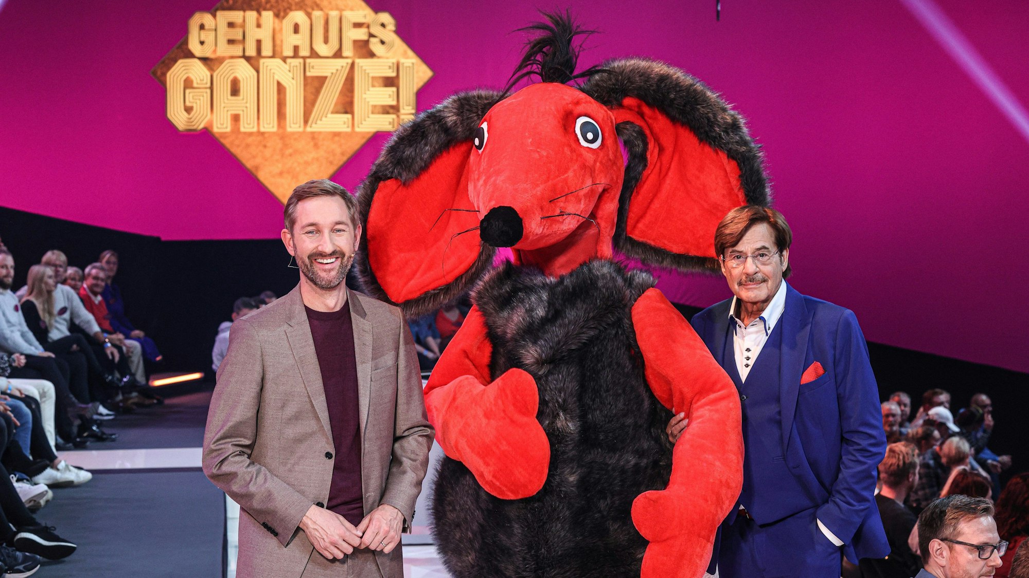 Daniel Boschmann (l-r), Zonk, eine menschengroße rote Maus, und Jörg Draeger in der 1. Staffel der Neuauflage der Kultshow «Geh aufs Ganze!», die auf Sat.1 ausgestrahlt wurde. Sie stehen vor einem goldenen Schild auf dem „Geh aufs Ganze!“ steht und lächeln in die Kamera.