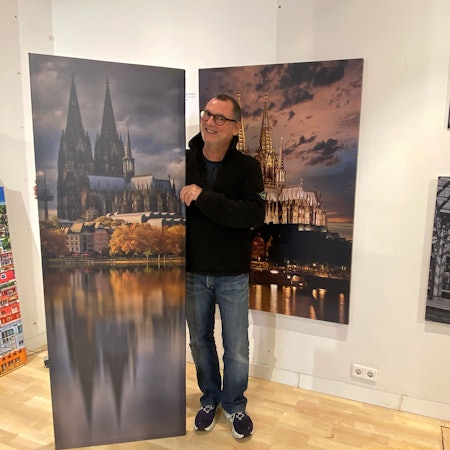 Ein Mann in einer schwarzen Jacke steht rechts rechts vor dem Bild in einer Galerie. Das Gemälde zeigt den Kölner Dom mit seiner Spiegelung im Wasser