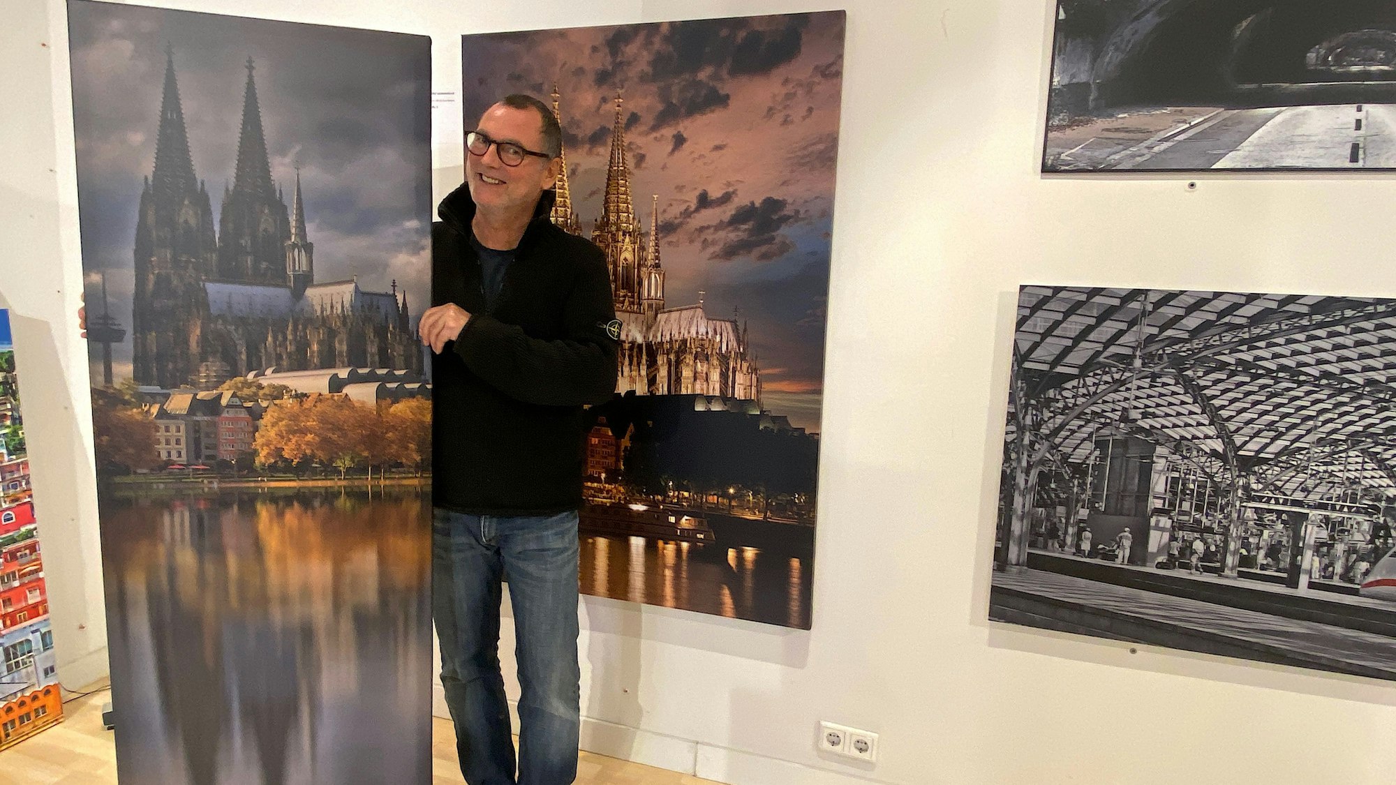 Ein Mann in einer schwarzen Jacke steht rechts rechts vor dem Bild in einer Galerie. Das Gemälde zeigt den Kölner Dom mit seiner Spiegelung im Wasser
