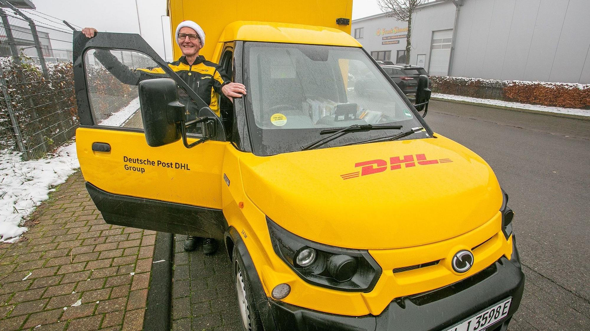 Ein Mann mit Mütze und einem Deutsche Post-Outfit steht in der Türe des gelben DHL-Transporters. Durch die Scheibe sieht man das Lenkrad auf der rechten Seite
