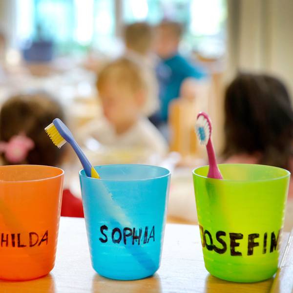 Zahnbürsten und Zahnputzbecher mit den Vornamen der Kinder stehen in einer Kindertagesstätte auf einem Tisch.