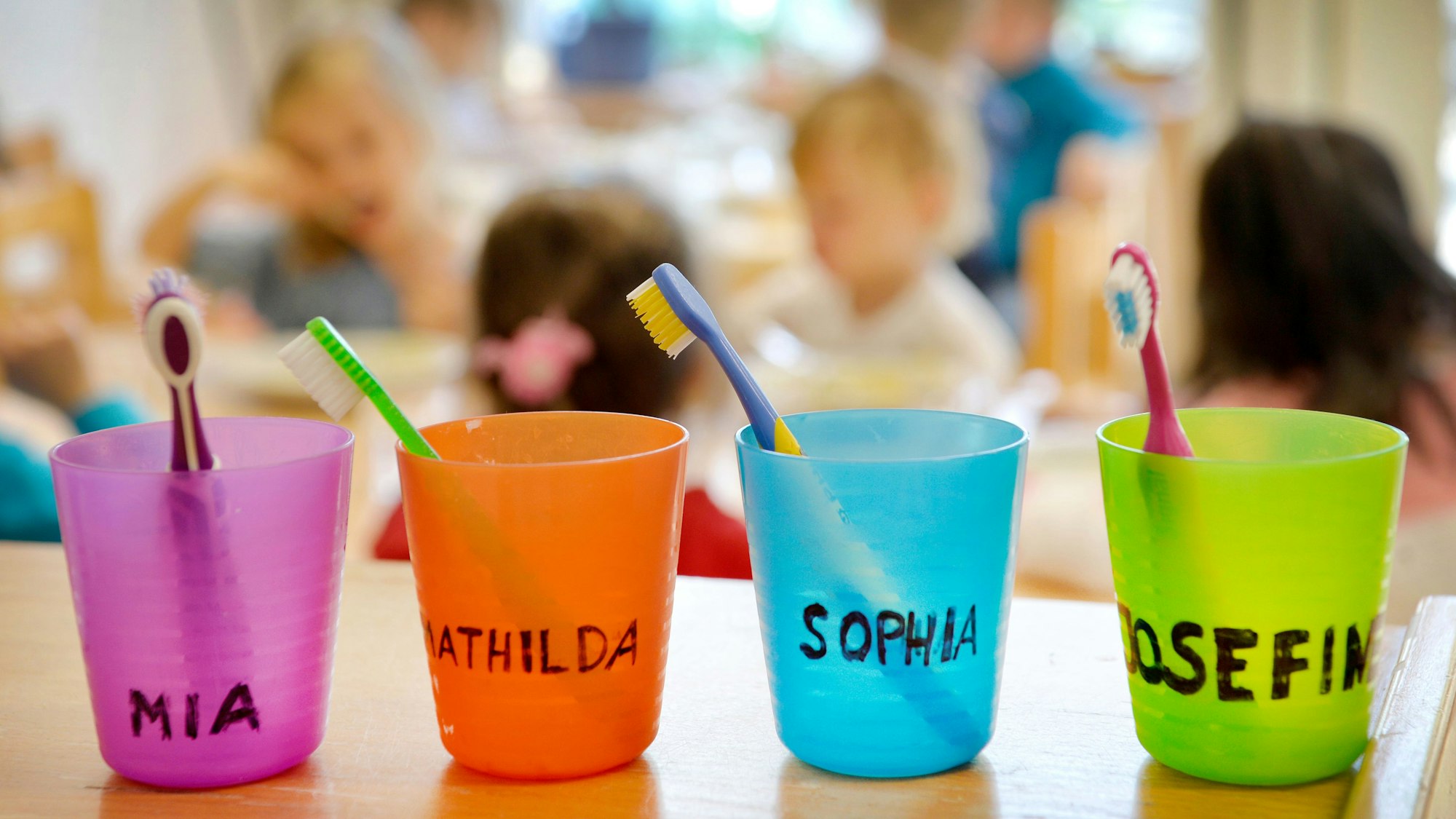 Zahnbürsten und Zahnputzbecher mit den Vornamen der Kinder stehen in einer Kindertagesstätte auf einem Tisch.