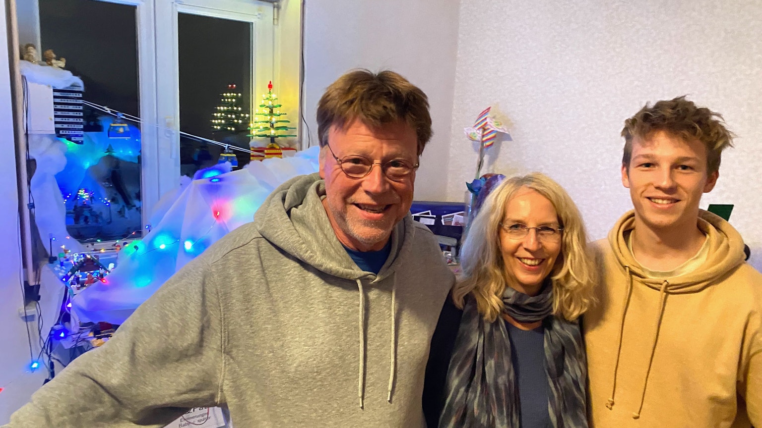 Ein Mann und eine Frau und ein jüngerer Mann, Eltern und Sohn, stehen nebeneinander, im Hintergrund sind mit Lichterketten beleuchtete Lego-Aufbauten zu sehen.
