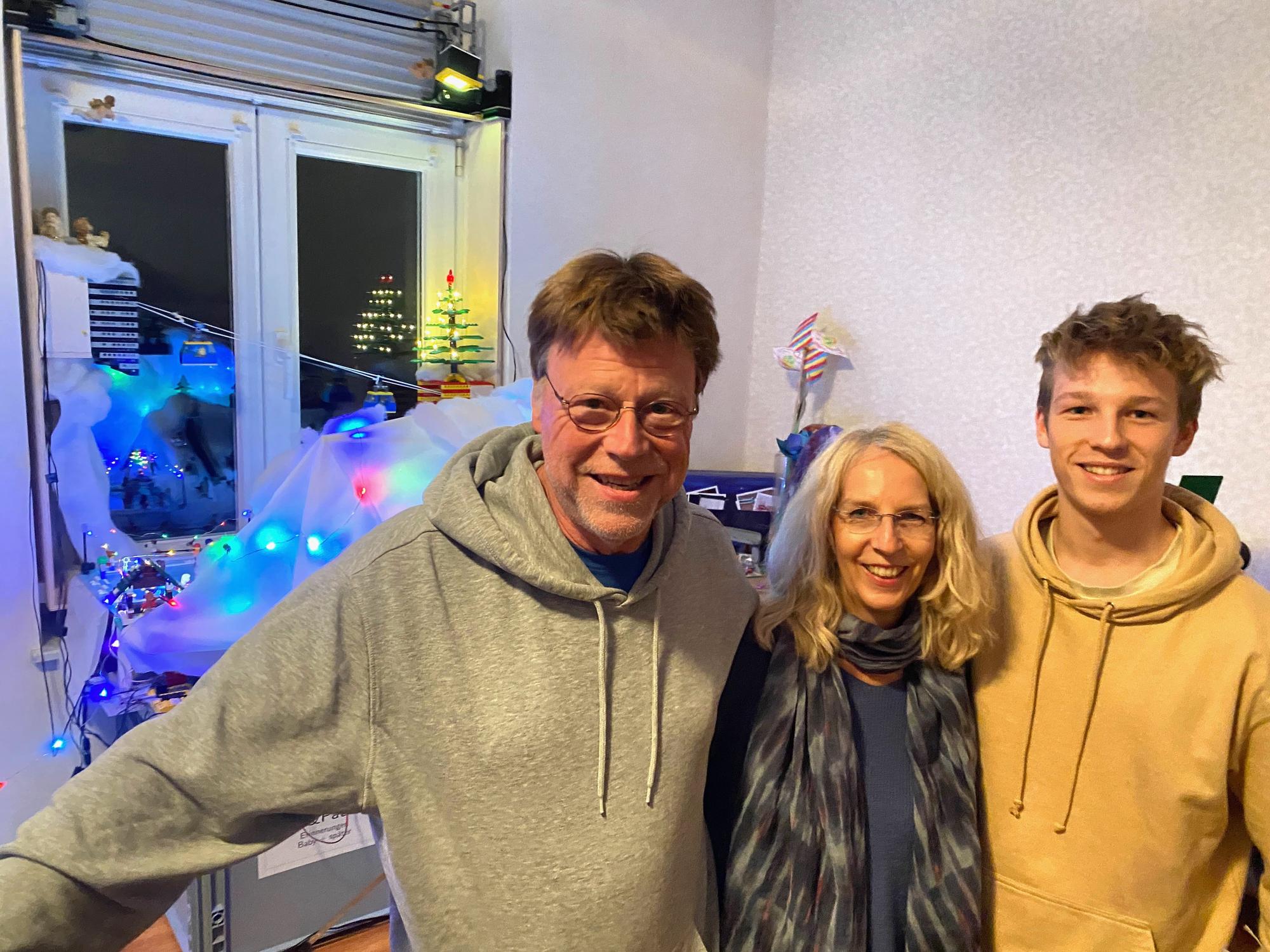Ein Mann und eine Frau und ein jüngerer Mann, Eltern und Sohn, stehen nebeneinander, im Hintergrund sind mit Lichterketten beleuchtete Lego-Aufbauten zu sehen.