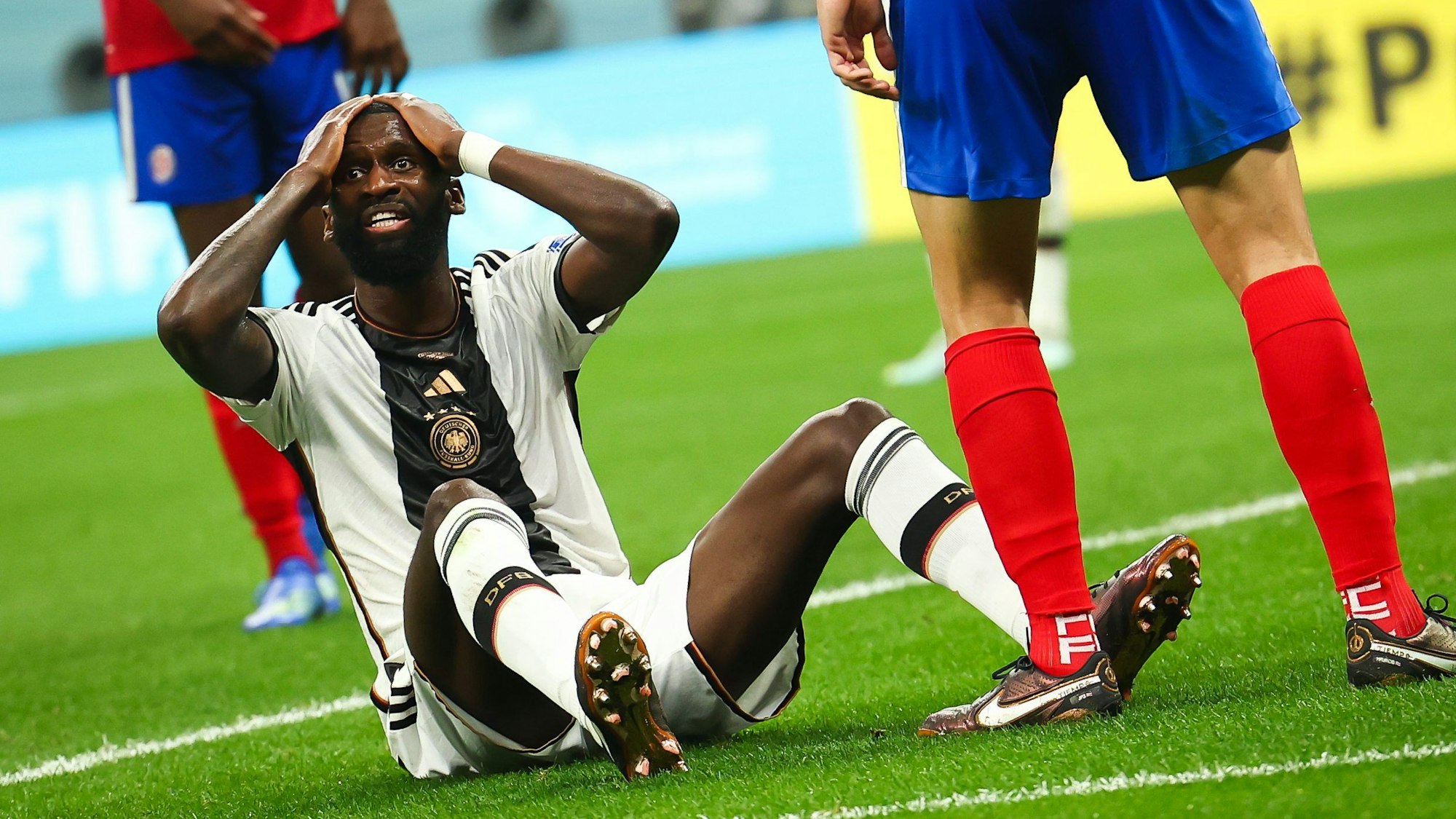 Der deutsche Nationalspieler Antonio Rüdiger sitzt auf dem Rasen während des WM-Spiels gegen Costa Rica. Er fasst sich an den Kopf. Außerdem sind die Beine von zwei costa-ricanischen Nationalspielern zu sehen.