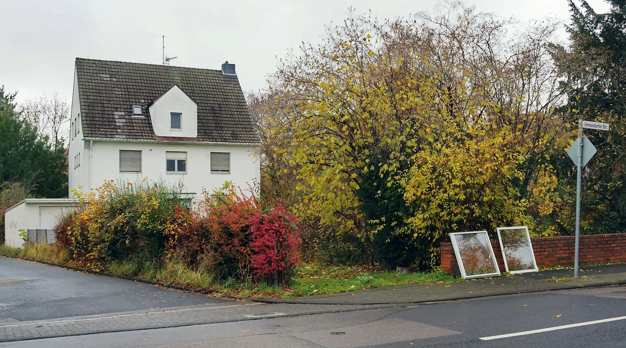Ein weißes Haus mit geschlossenen Rollläden und einem dunklen Dach ist zu sehen. Davor ist ein verwilderter Garten mit Sträuchern und Bäumen. An der Mauer zur Straße lehnen zwei kaputte Fensterrahmen.