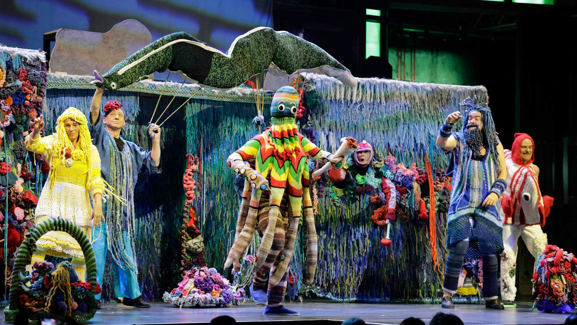 Das Bild zeigt das Unterwasser-Bühnenbild des Sketches „Wolle“. Das Ensemble der Stunksitzung stellt zum Beispiel einen Oktopus mit Reggae-Verkleidung, die Loreley, Vater Rhein oder einen Fisch namens Doktor Doktorfisch das. Alle Kostüme sind aus Wolle gestrickt.