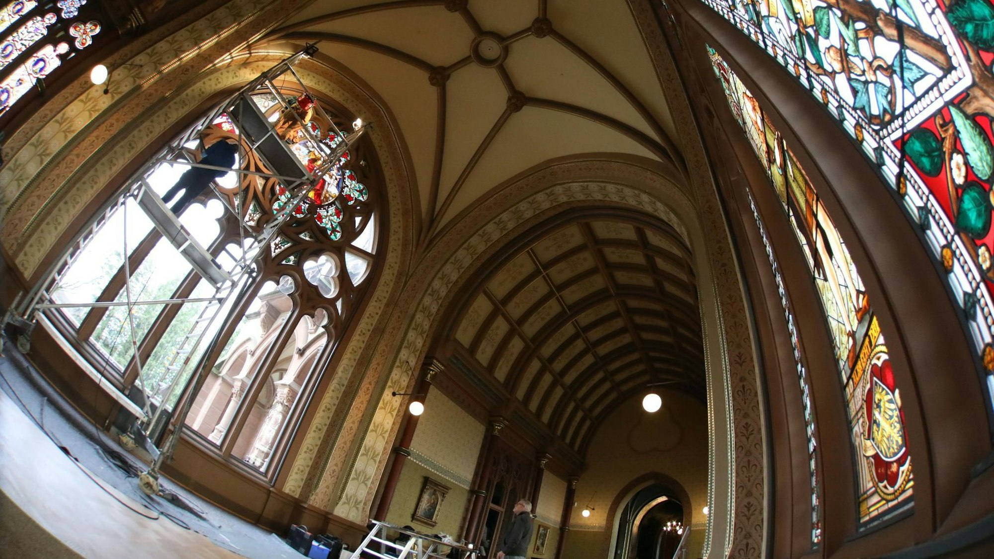 In der großen und hohen Kunsthalle von Schloss Drachenburg sind rechts die schon rekonstruierten Buntglasscheiben zu sehen. Auf der linken Seite setzen die Experten von einem Gerüst aus die letzten Scheiben ein.