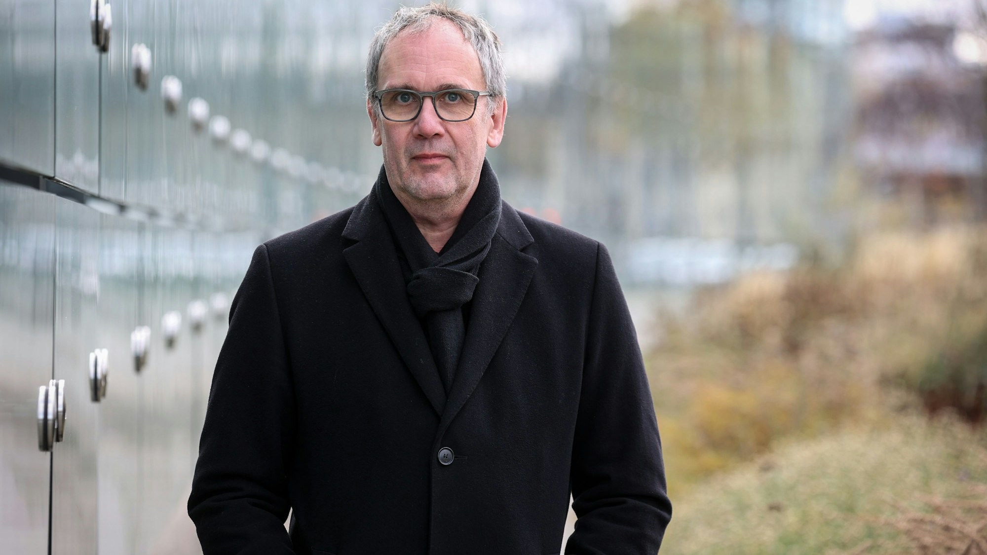 Bestseller-Autor Volker Kutscher steht neben einer Glasfassade und schaut in die Kamera. Er trägt einen schwarzen Mantel und Schal.