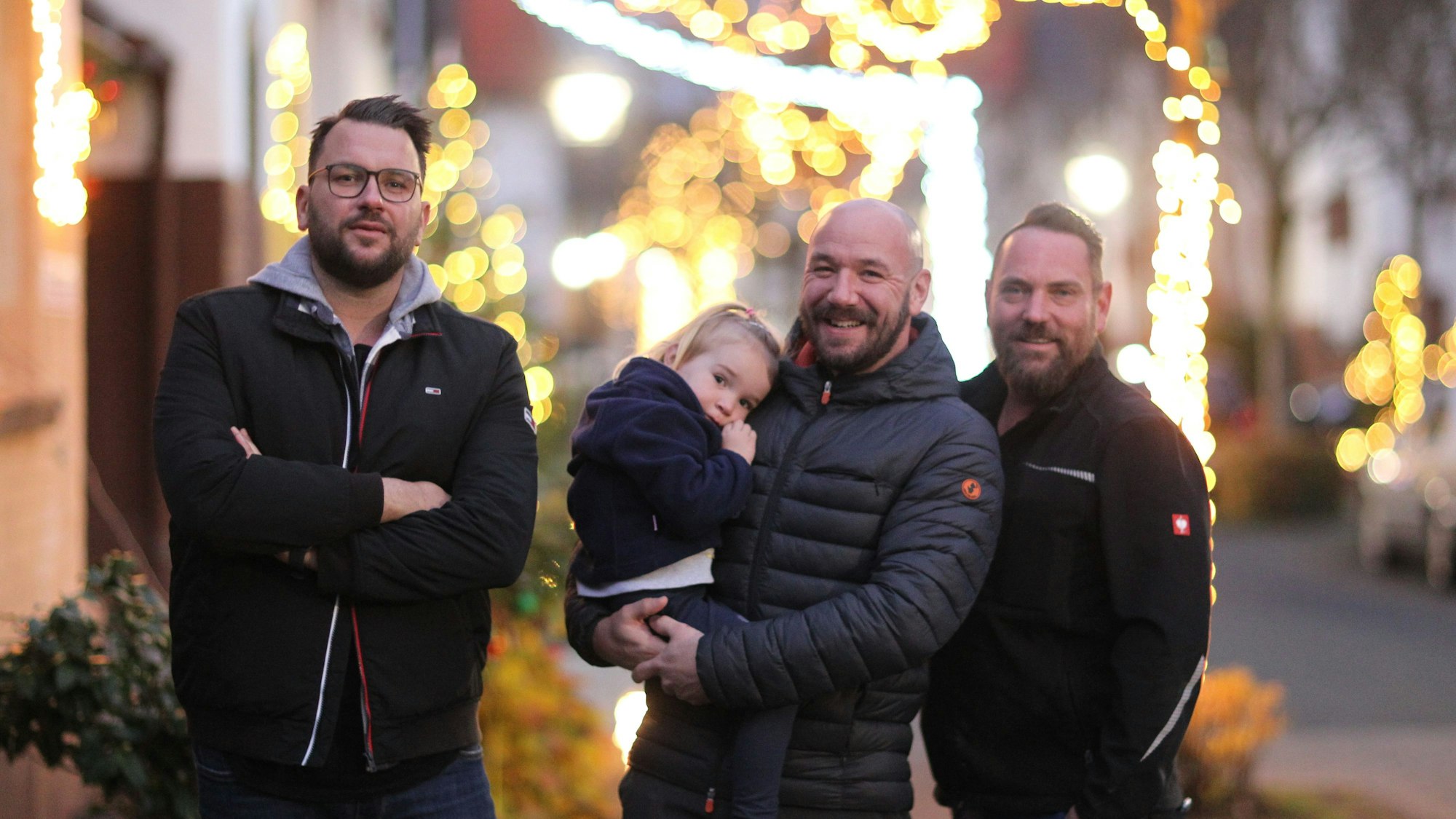 Die Initiatoren der Weihnachtsstraße (von links) Patrick Schübl, Daniel Thimm (mit Töchterchen Flora) und Thorsten Simmerkus stehen in der Dämmerung vor dem Lichterglanz an Bäumen und Vorgärte.