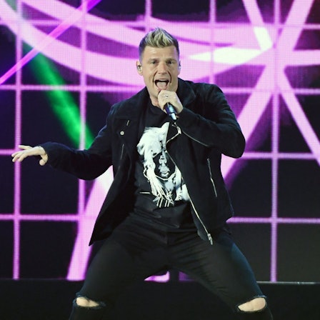Backstreet-Boys-Sänger Nick Carter hält ein Mikrofon in der Hand und singt auf einer Bühne vor pinkem-schwarzem Hintergrund.