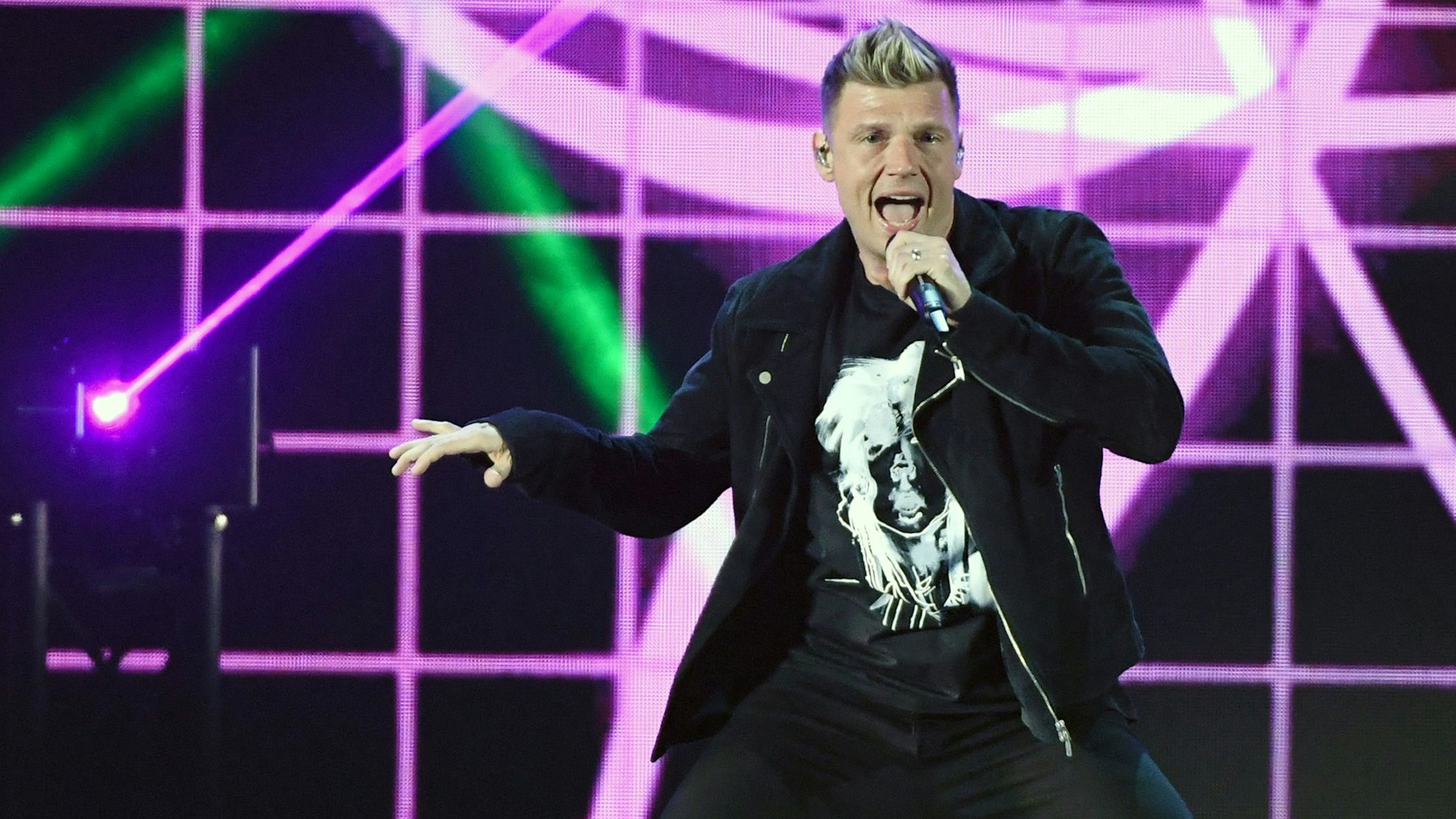 Backstreet-Boys-Sänger Nick Carter hält ein Mikrofon in der Hand und singt auf einer Bühne vor pinkem-schwarzem Hintergrund.