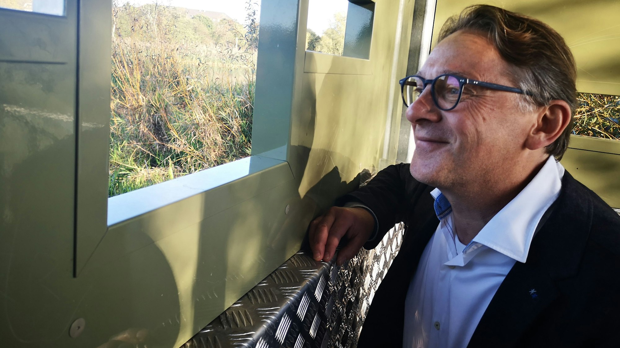 Auf dem Foto ist ein Mann mit Brille zu sehen, der aus einem Fenster blickt.