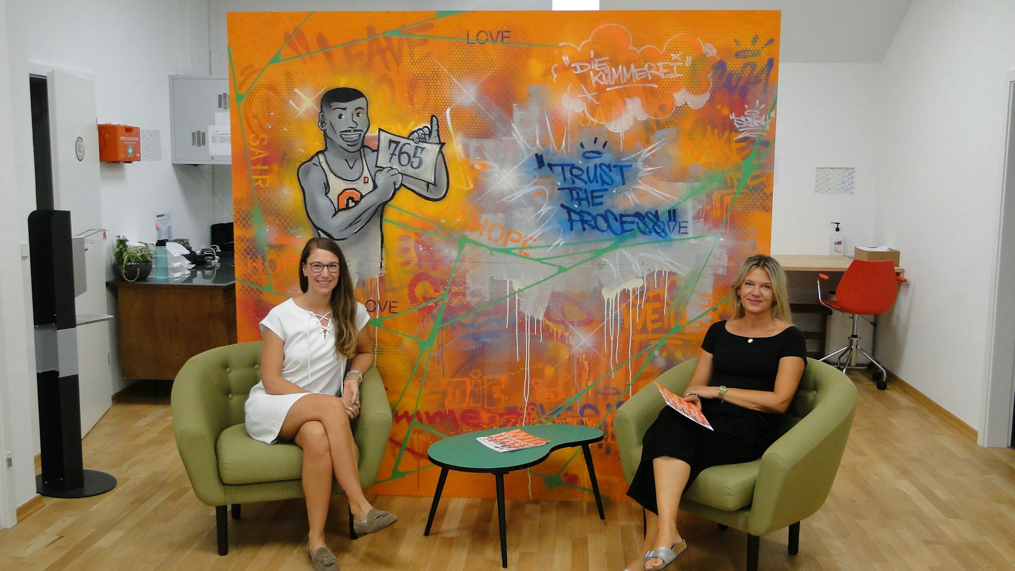Sandra Sändker von der AOK Rheinland/Hamburg und die Leiterin der Kümmerei, Birgit Skimutis, sitzen vor einem bunten großen Bild auf Sesseln.