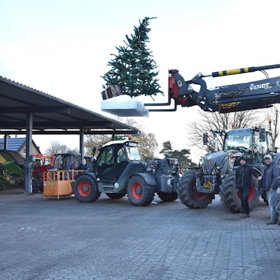 Mehrere Traktoren stehen auf einem Hof, zwei Männer in der Mitte. Ein Traktor hebt einen Tannenbaum.