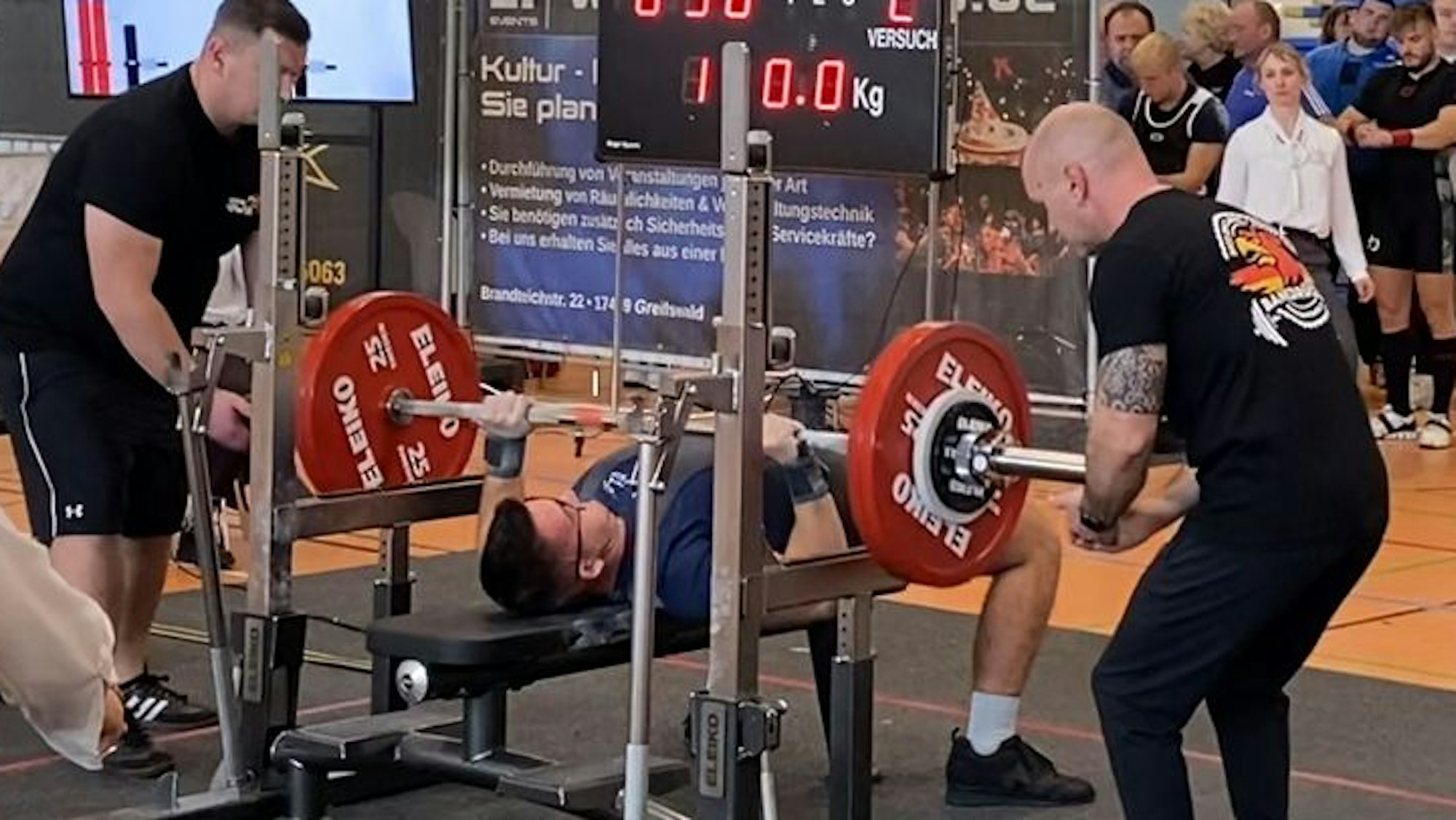 Der Athlet aus Pulheim liegt bei den Deutschen Meisterschaften in Greifswald auf der Bank, zwei Helfer assistieren ihm.