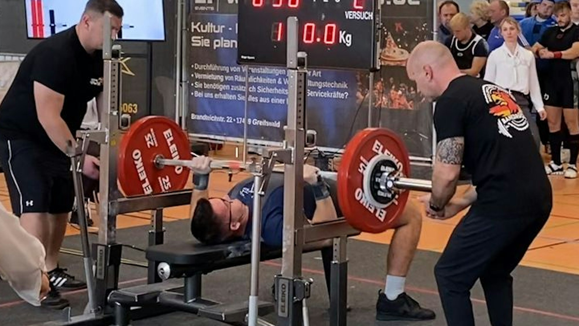 Der Athlet aus Pulheim liegt bei den Deutschen Meisterschaften in Greifswald auf der Bank, zwei Helfer assistieren ihm.