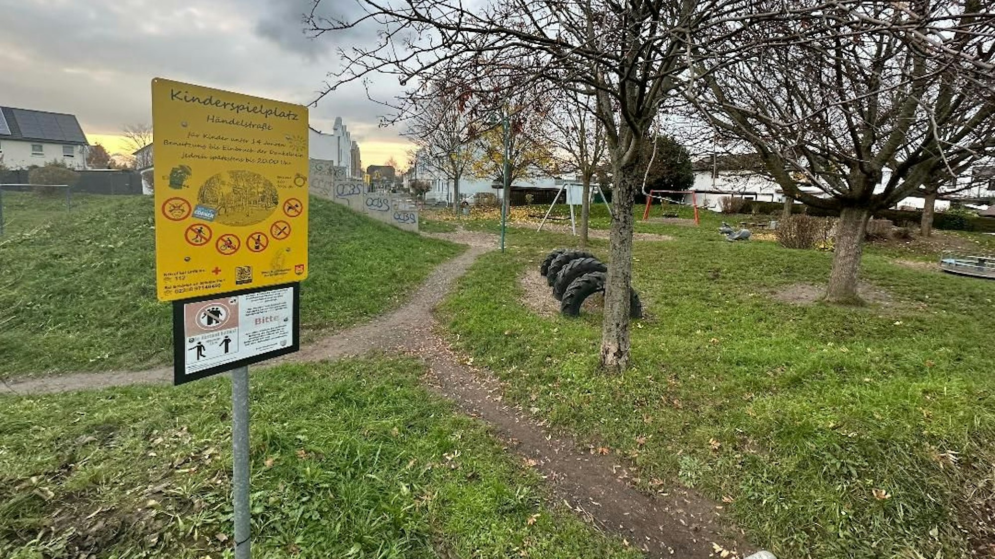 Ein leerer, heruntergekommener Spielplatz in Niederkassel. Im Vordergrund ist ein gelbes Schild, auf dem "Kinderspielplatz Händelstraße" steht. Im Hintergrund sind Spielgeräte wie Schaukeln, Reifen und Wippen zu sehen.