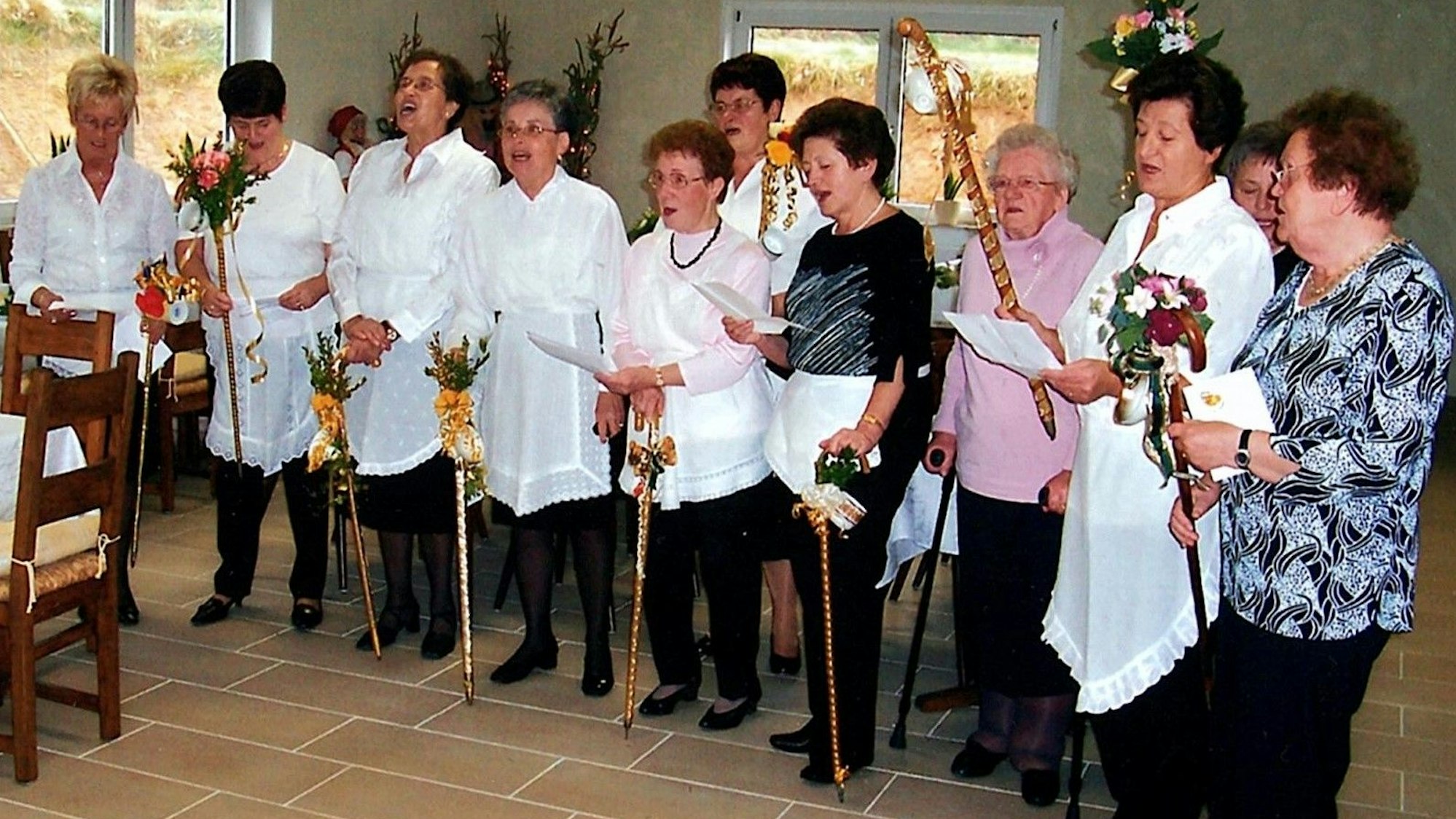 Ein altes Foto zeigt die Kaffeetanten bei einem Auftritt auf einer Feier in einem Saal. Die Frauen tragen Schürzen mit Spitze und halten Stöcke mit Kaffeetassen.