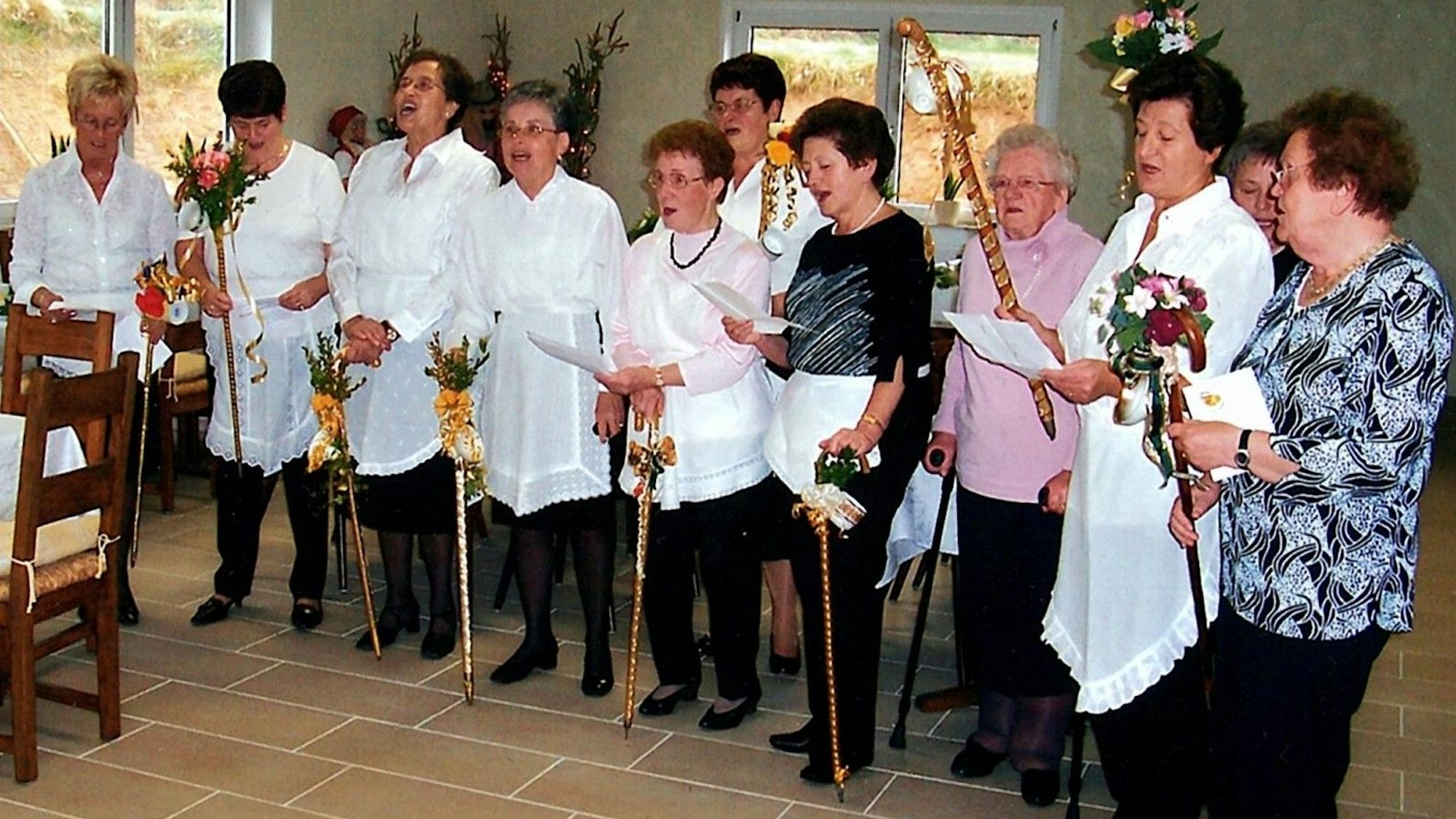 Ein altes Foto zeigt die Kaffeetanten bei einem Auftritt auf einer Feier in einem Saal. Die Frauen tragen Schürzen mit Spitze und halten Stöcke mit Kaffeetassen.