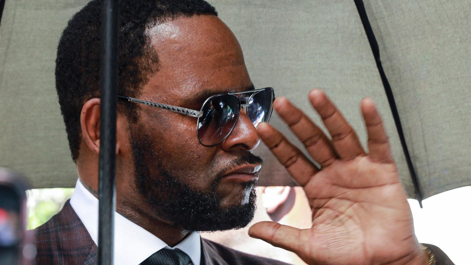 R. Kelly mit einer Sonnenbrille unter einem Regenschirm hebt die Hand in Richtung der Kamera.