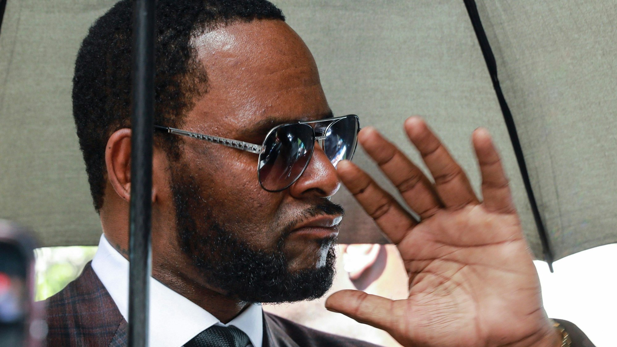 R. Kelly mit einer Sonnenbrille unter einem Regenschirm hebt die Hand in Richtung der Kamera.