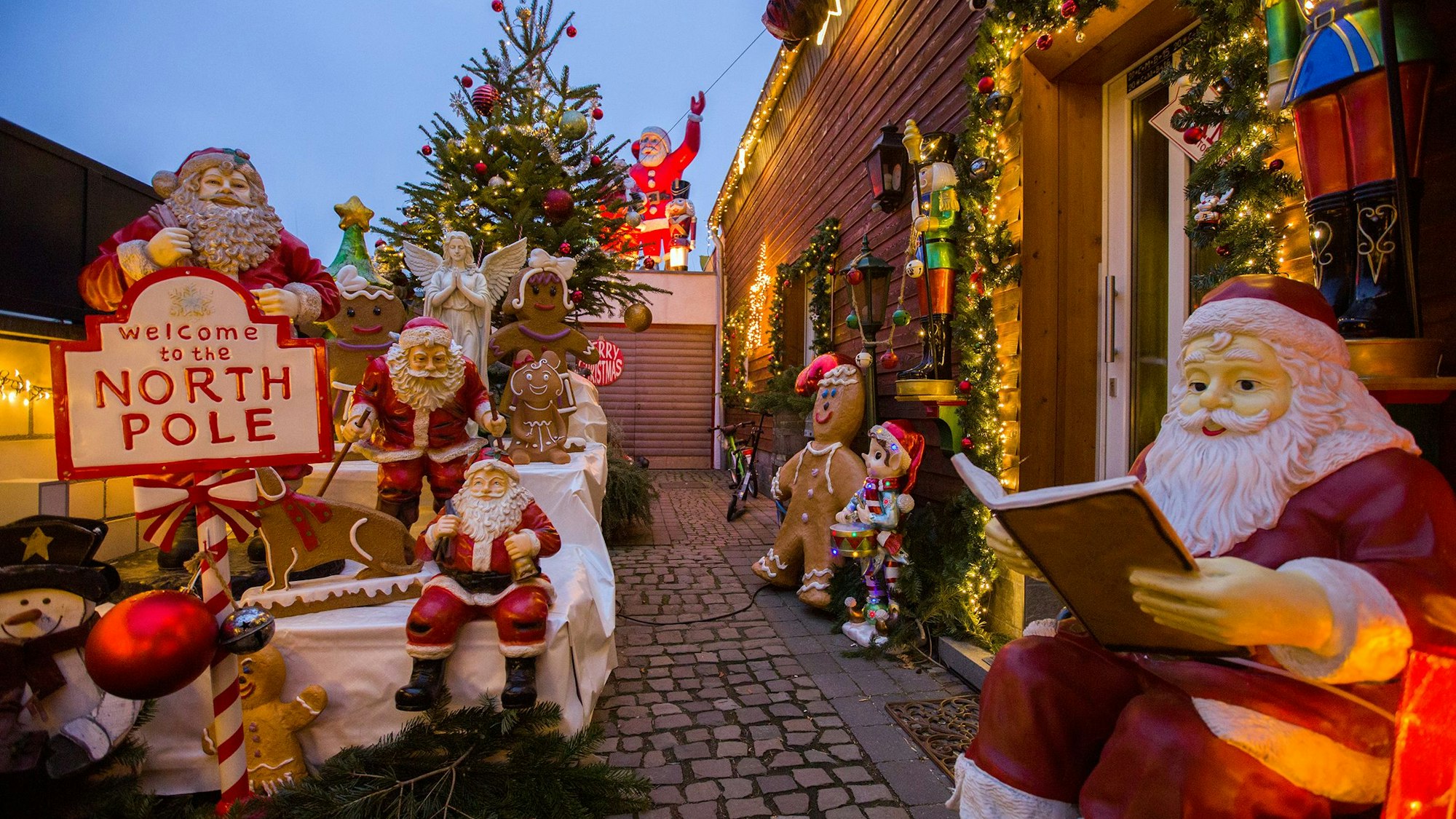 Mehrere Weihnachtsmänner, aber auch ein Engel und Lebkuchenfiguren stehen vor einem holzgetäfelten Haus. Auch ein geschmückter Weihnachtsbaum und ein Nikolaus auf einem Garagendach sind zu sehen.