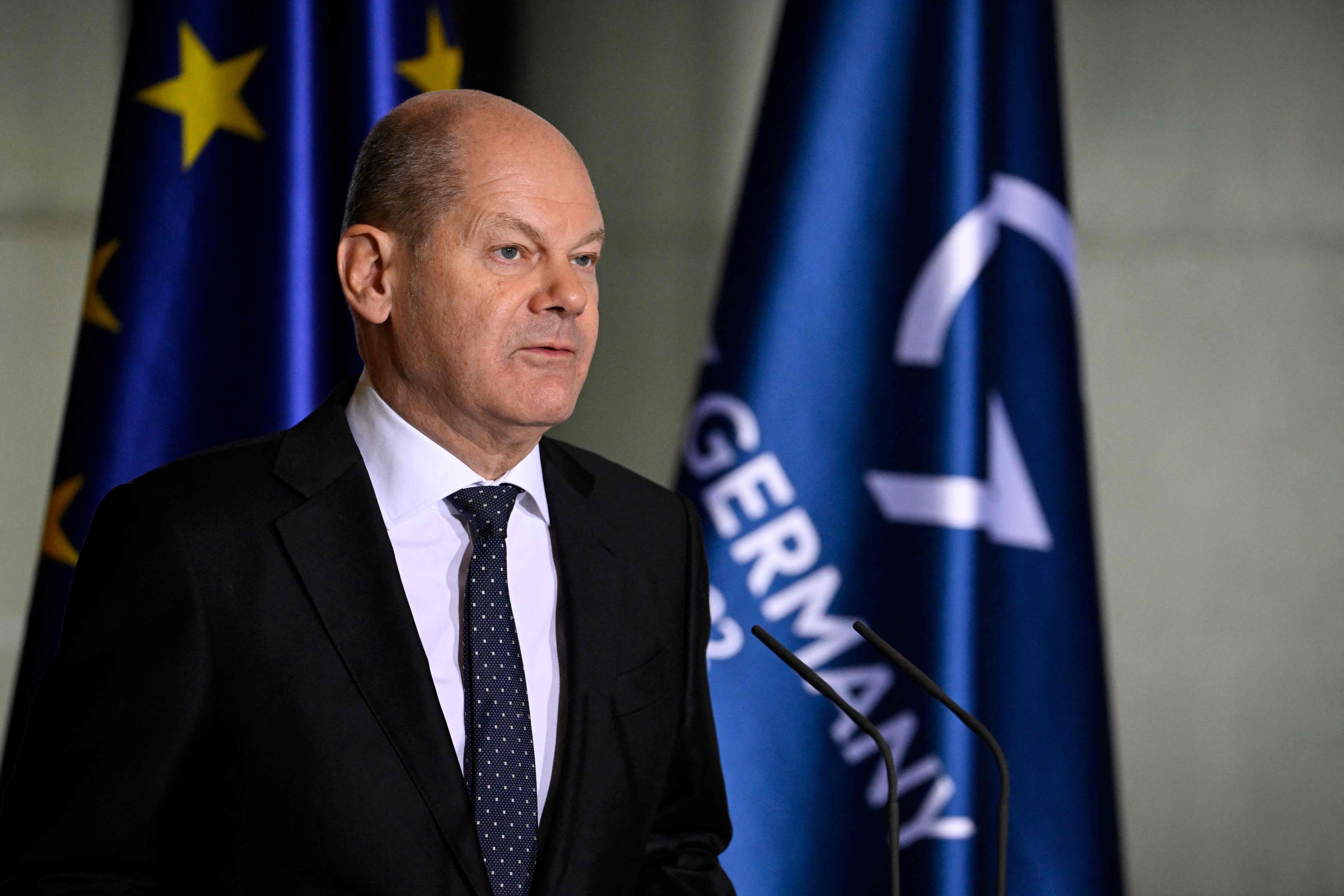 Olaf Scholz im Vordergrund steht vor Mikros und redet. Im Hintergrund sind die europäische Flagge und die der G7 zu sehen.