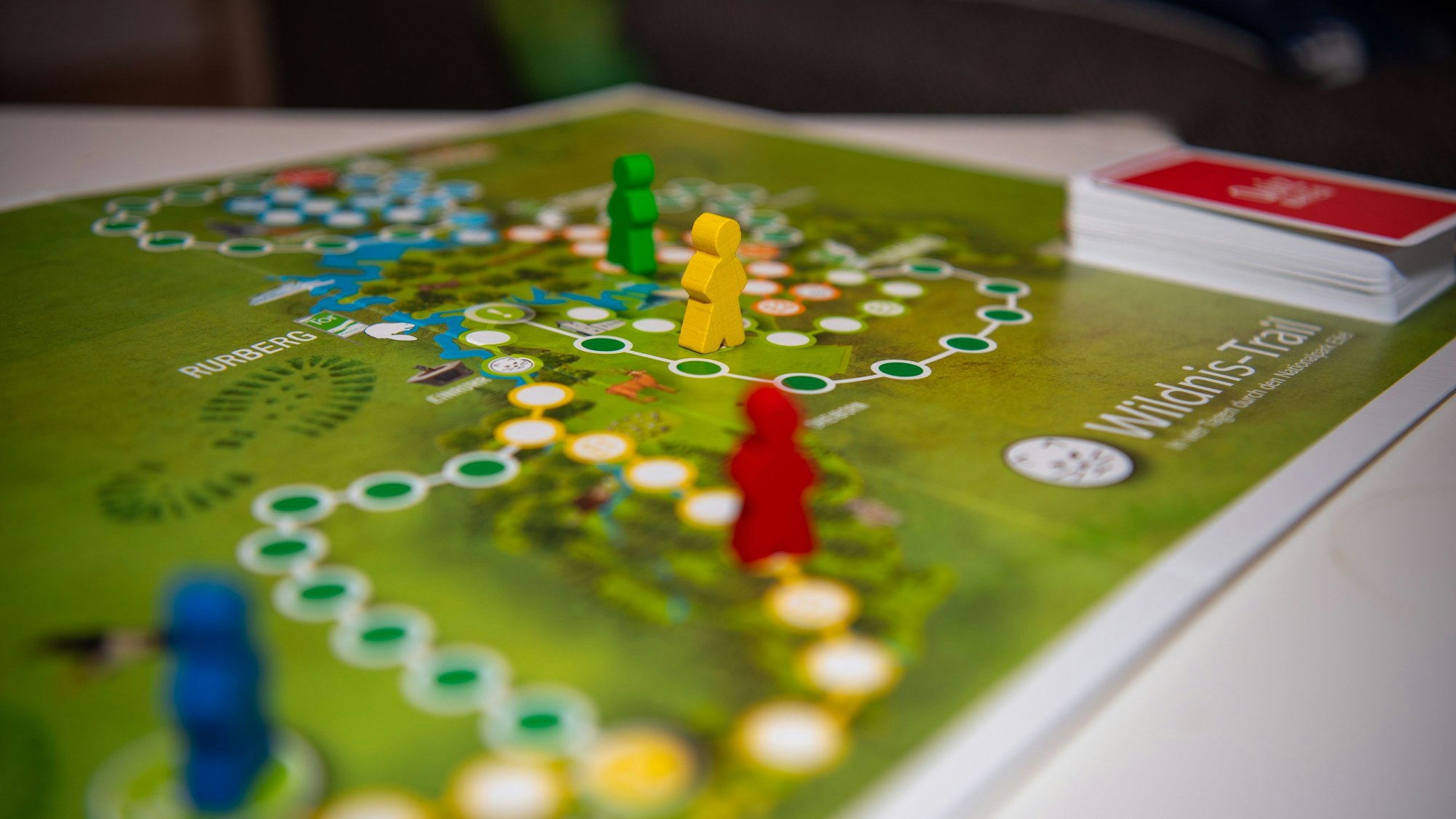 Das Bild zeigt ein Brettspiel mit vier bunten Figuren und einem Kartenstapel.