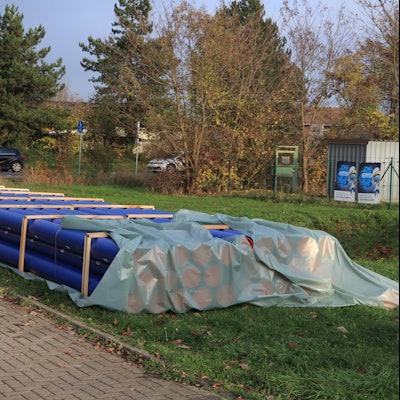 Die neuen blauen Abwasserrohre liegen bereits auf einer Wiese neben der Straße bereit.