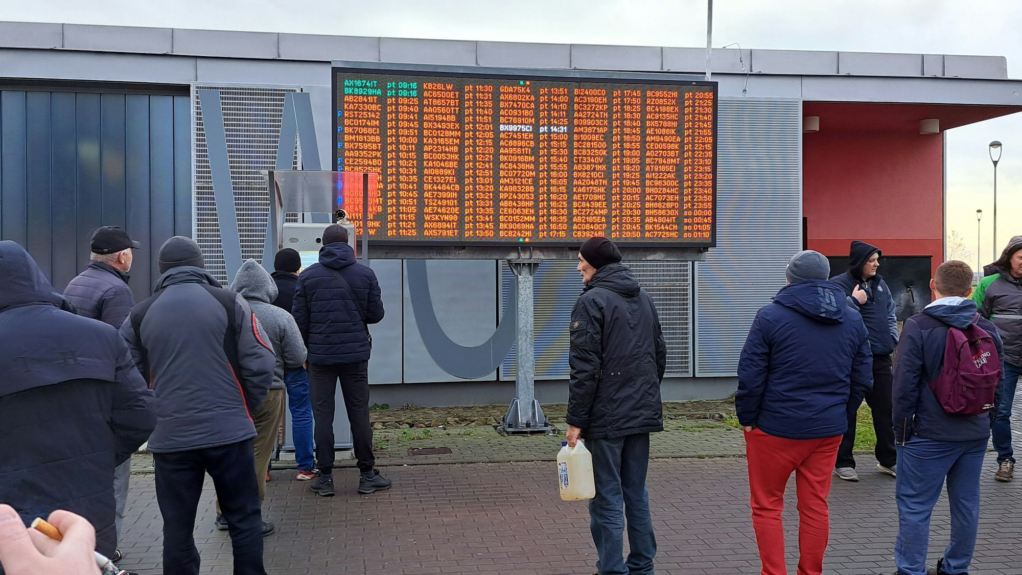 Menschen stehen vor einer großen Anzeigetafel im Freien.