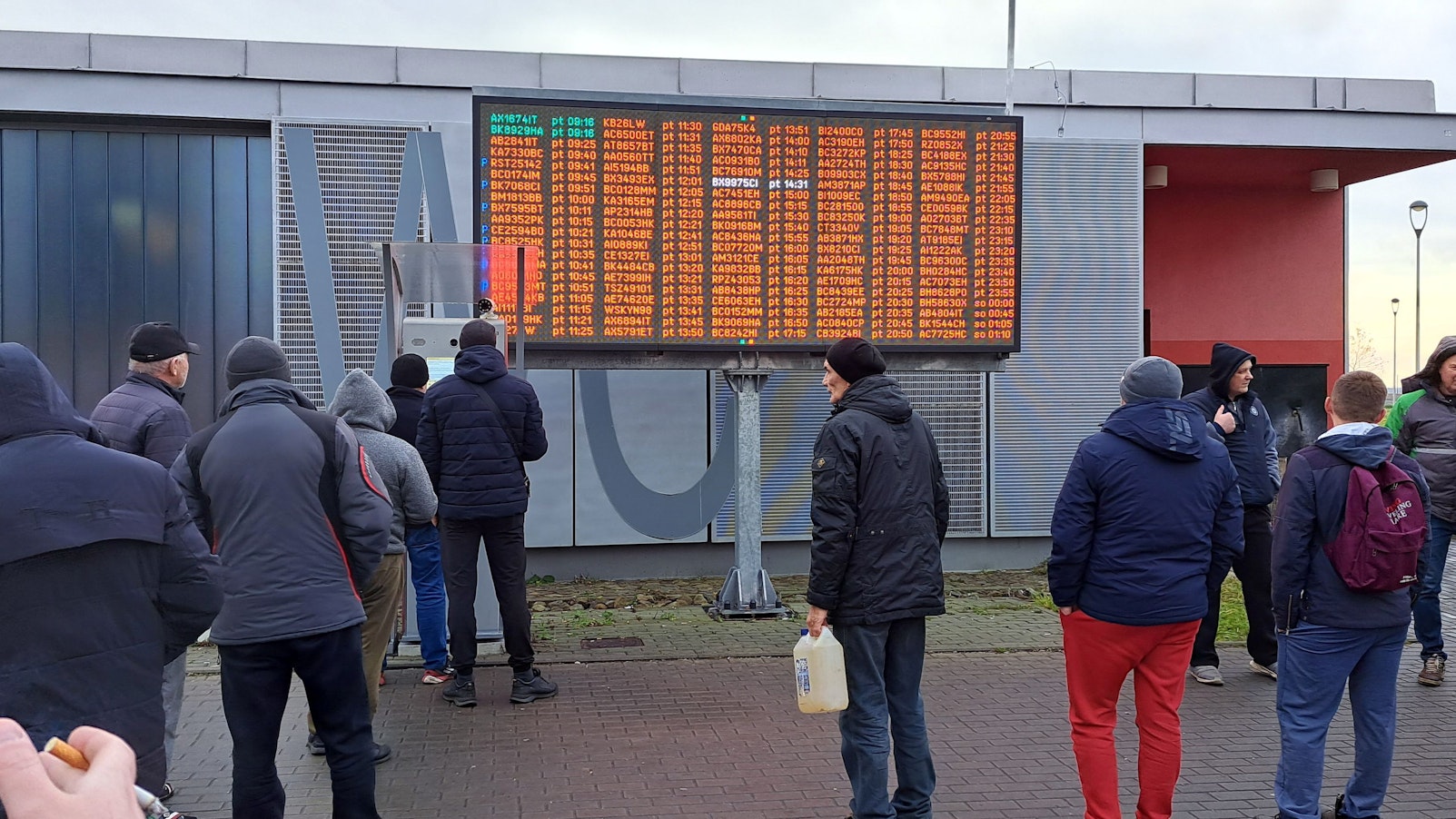 Menschen stehen vor einer großen Anzeigetafel im Freien.