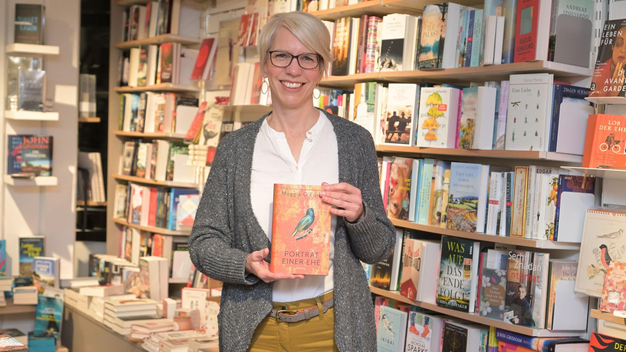 Andrea Etzweiler steht vor einem Bücherregal und hält „Porträt einer Ehe“ in der Hand.
