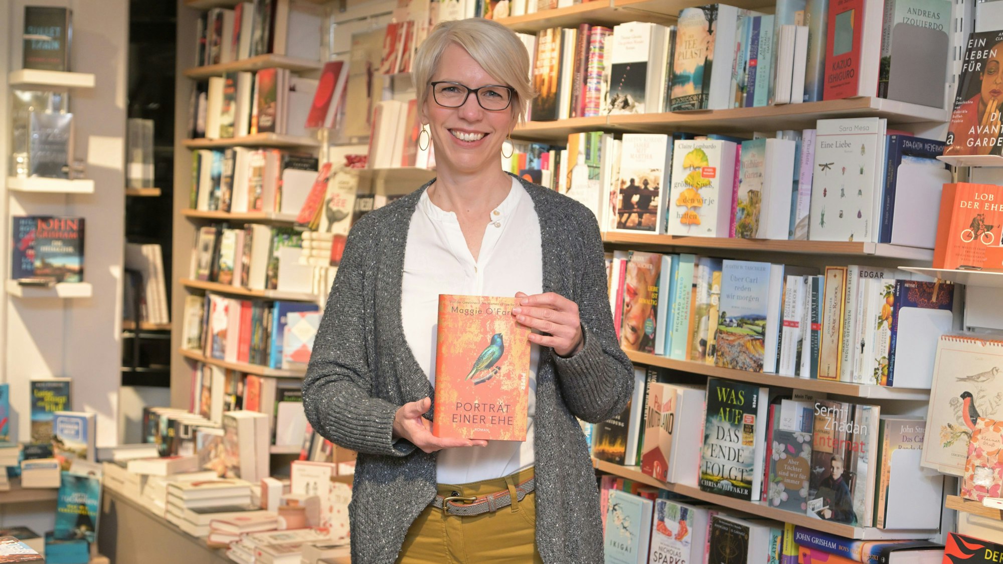 Andrea Etzweiler steht vor einem Bücherregal und hält „Porträt einer Ehe“ in der Hand.